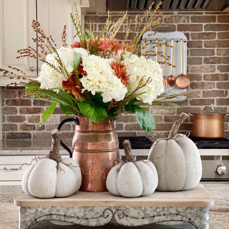 Three concrete pumpkins displayed on a riser on a kitchen island with a copper pitcher filled with Fall florals.
