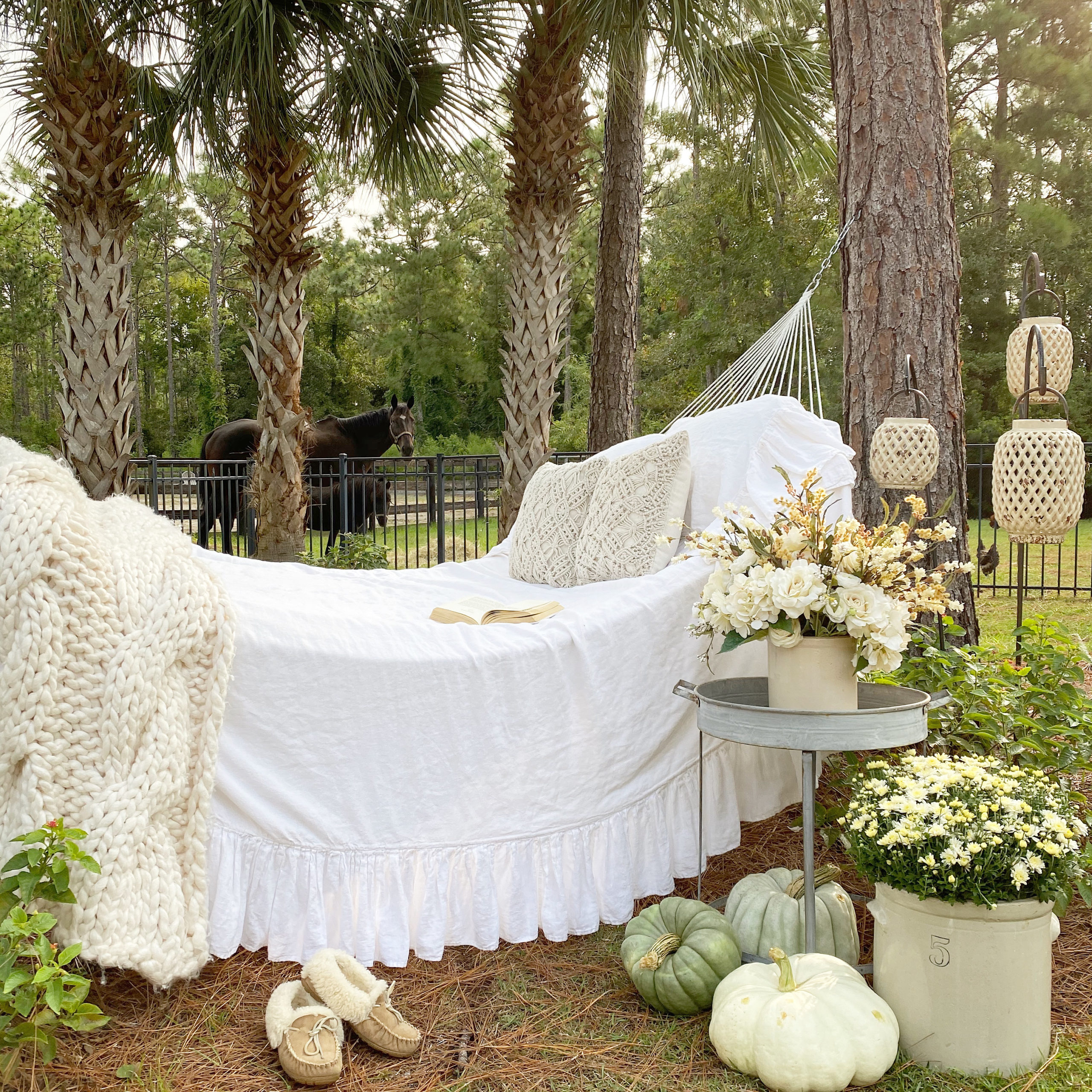 A cozy hammock set up for Fall with a ruffled white linen cover, a Fall floral arrangement on a metal table next to it and pumpkins and mums for decoration around it.