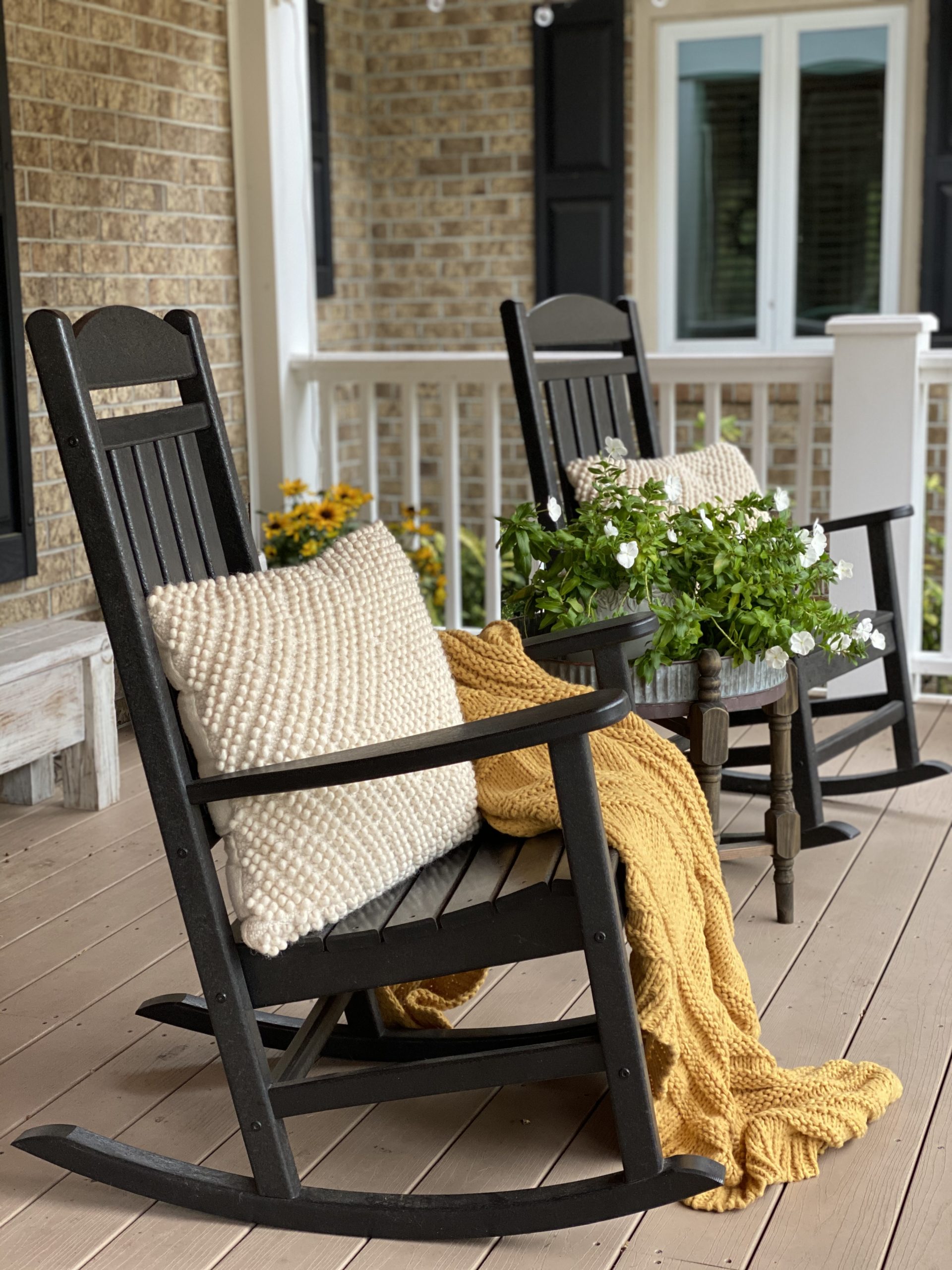 Front porch with black rocking chairs and a mustard yellow blanket, coat cream pillows and flowers.