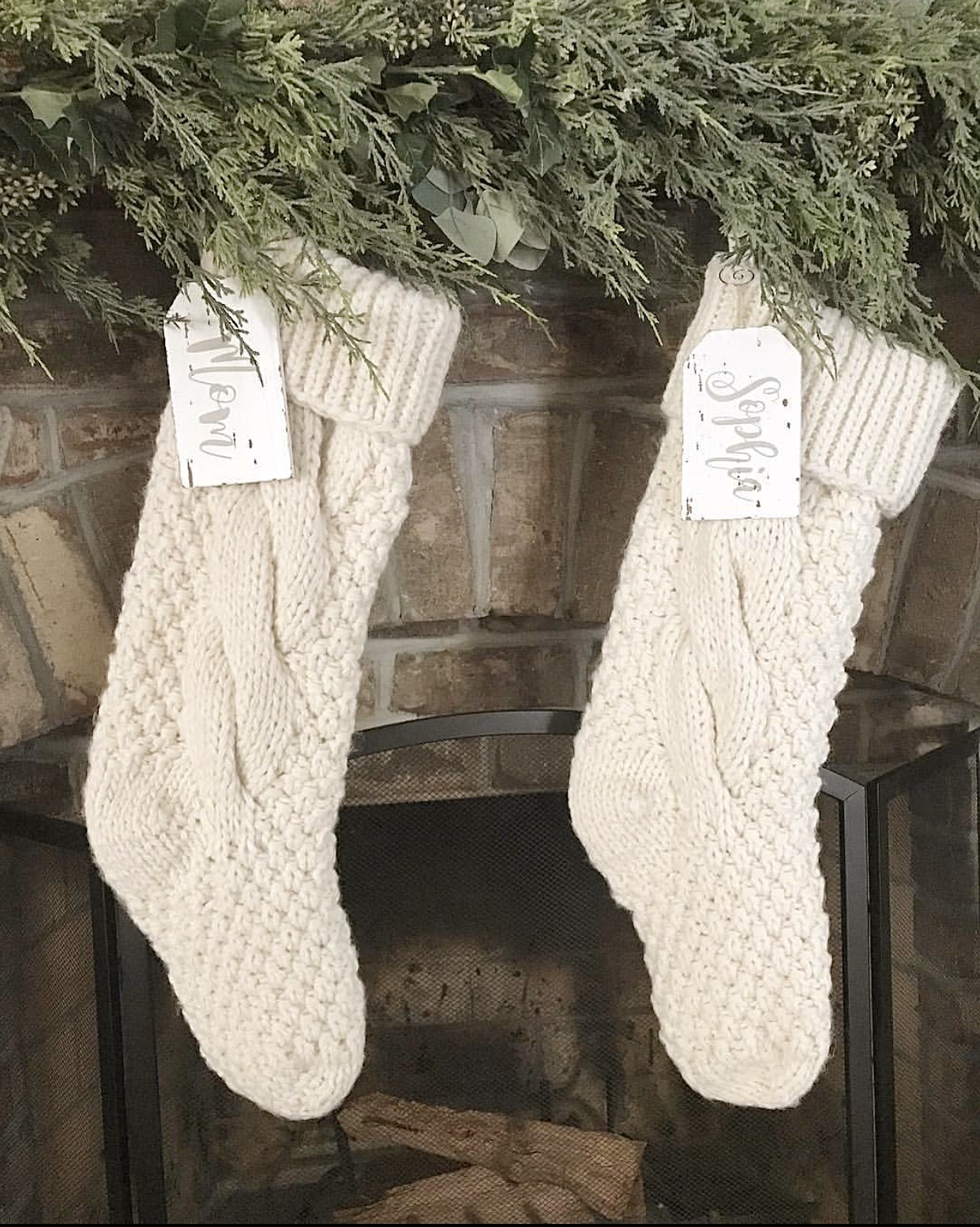 White knit stockings hung in front of a brick fireplace with garland. 