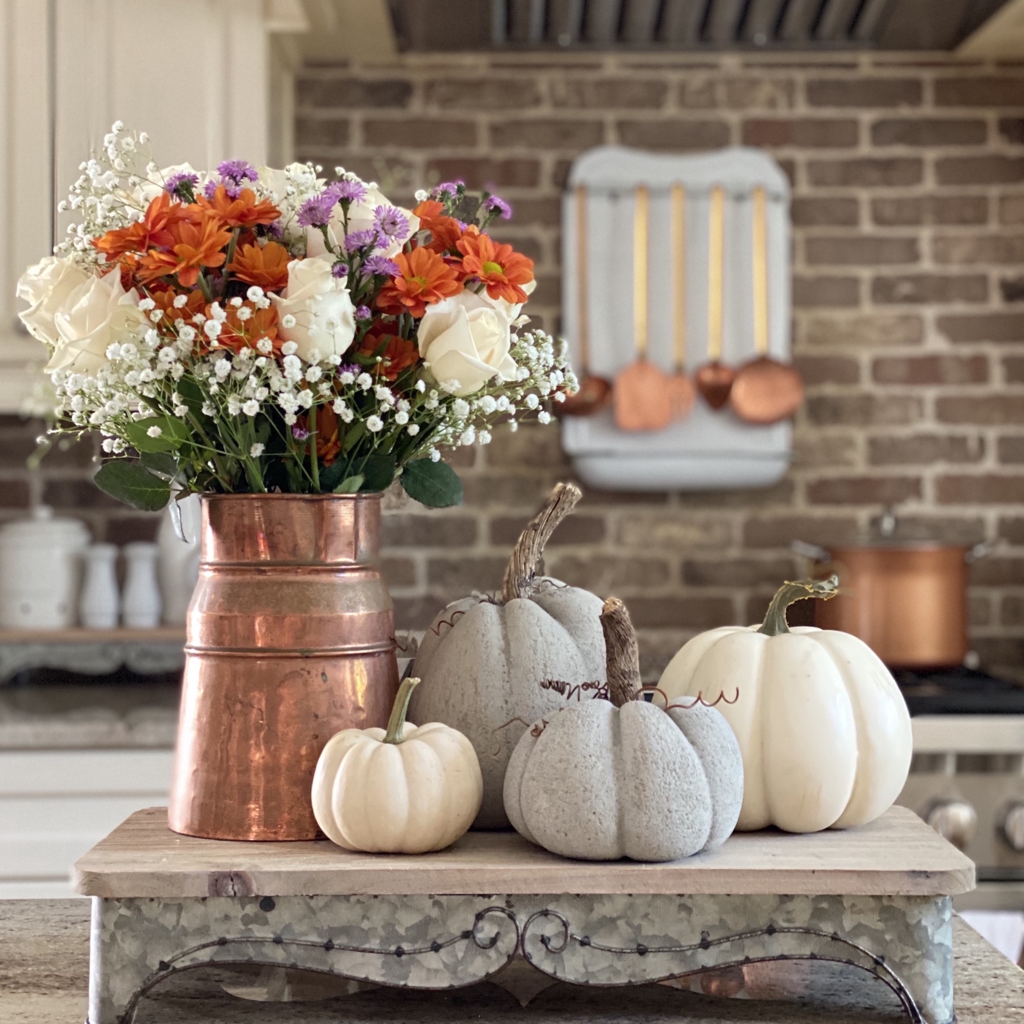 A copper pitcher on a riser with a fall floral arrangement in it, surrounded by pumpkins. In the background is a copper pot on the stove and and copper utensils hanging above it.
