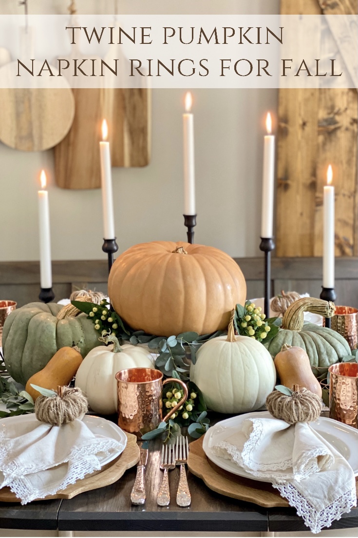Fall table set with white dishes and linen napkins with twine pumpkin rings around them. The centerpiece is made with lots of pumpkins and candles.