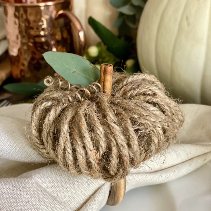 A linen napkin with a wood napkin ring around it and a jute twine pumpkin on top.
