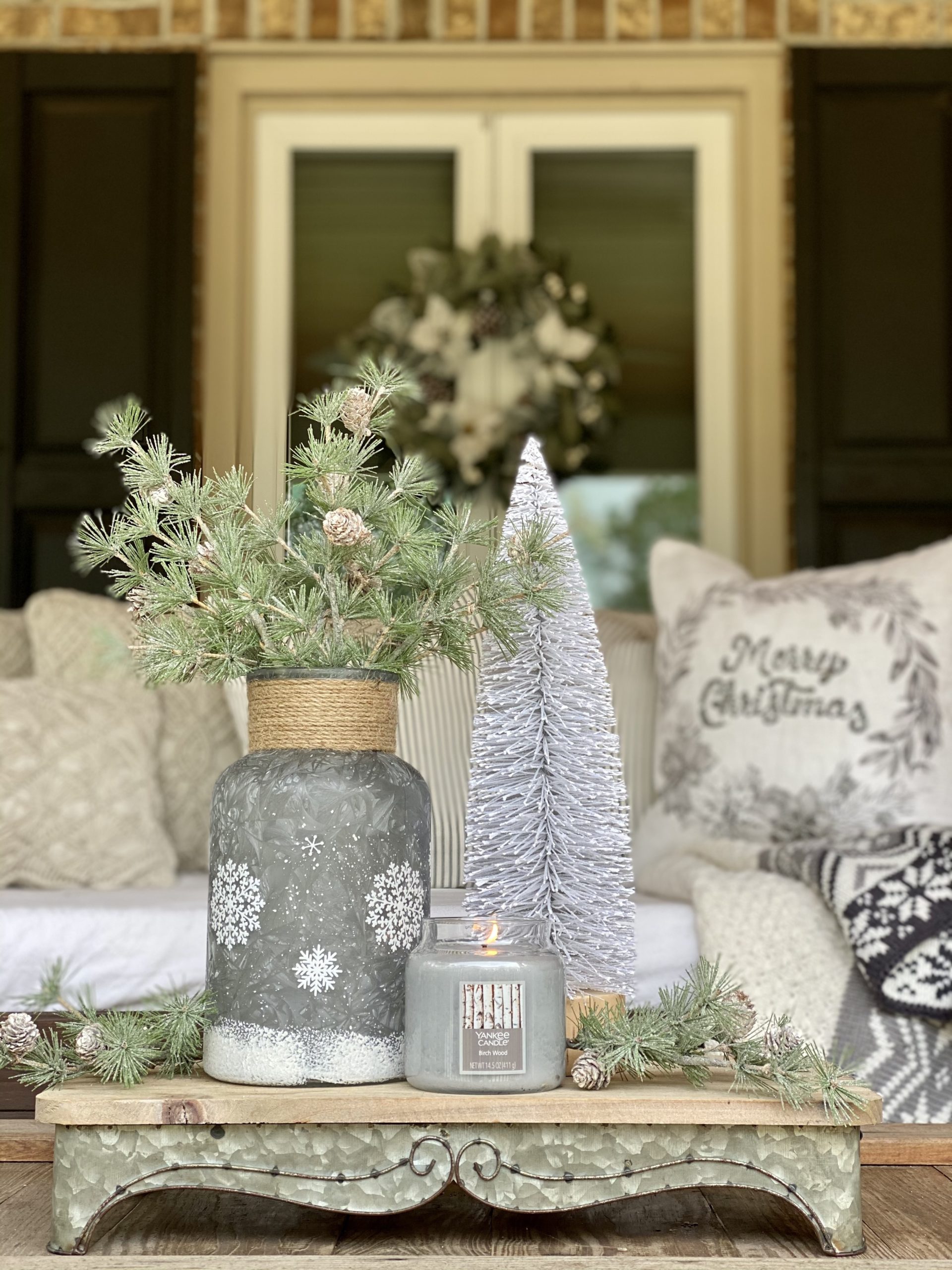 Coffee table Christmas decor in front of porch swing. Tray includes bottle brush tree, vase with greenery and a candle