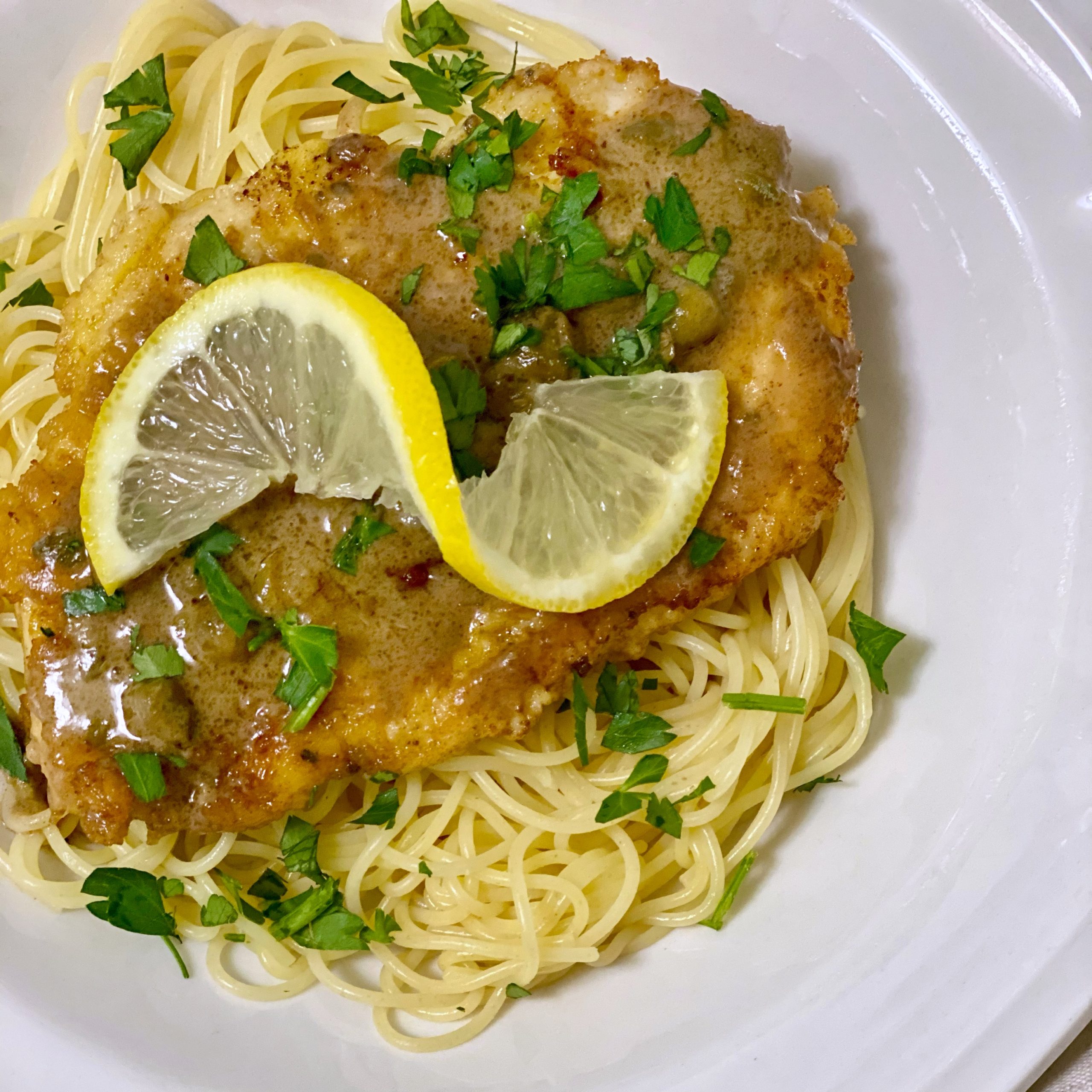Lemon Chicken with capers and parsley with a lemon for garnish on a bed of angel hair.