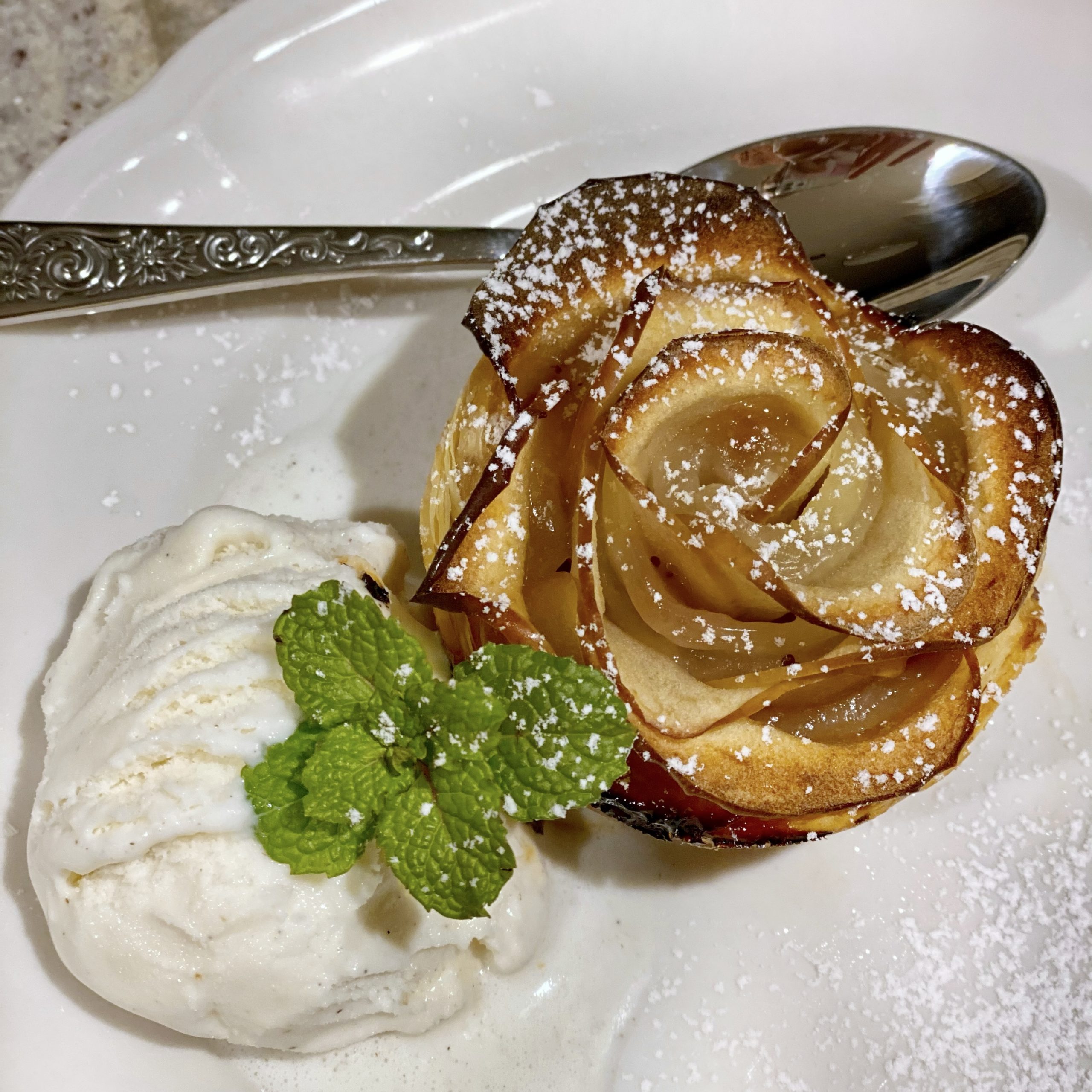 Baked Apple Rose on a white plate with a scoop of vanilla ice cream and a sprig of mint garnish.