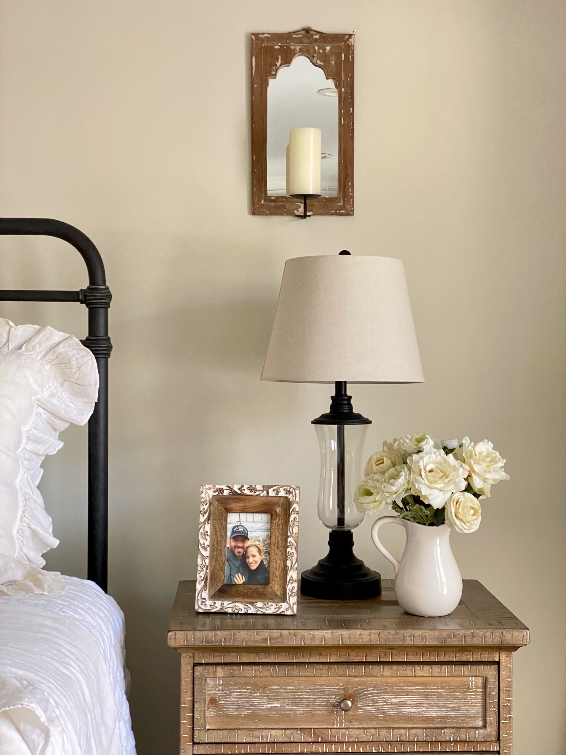 The perfect bedside essentials: bronze and glass table lamp, flowers in a pitcher and a picture frame on a wood nightstand next to the bed.