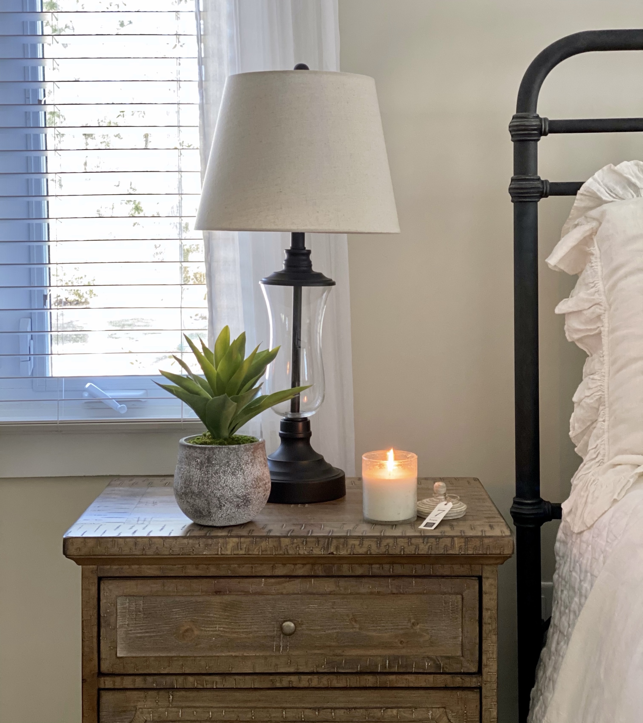 The perfect bedside essentials: a faux plant, candle, and bronze and glass lamp on a wood nightstand next to the bed.