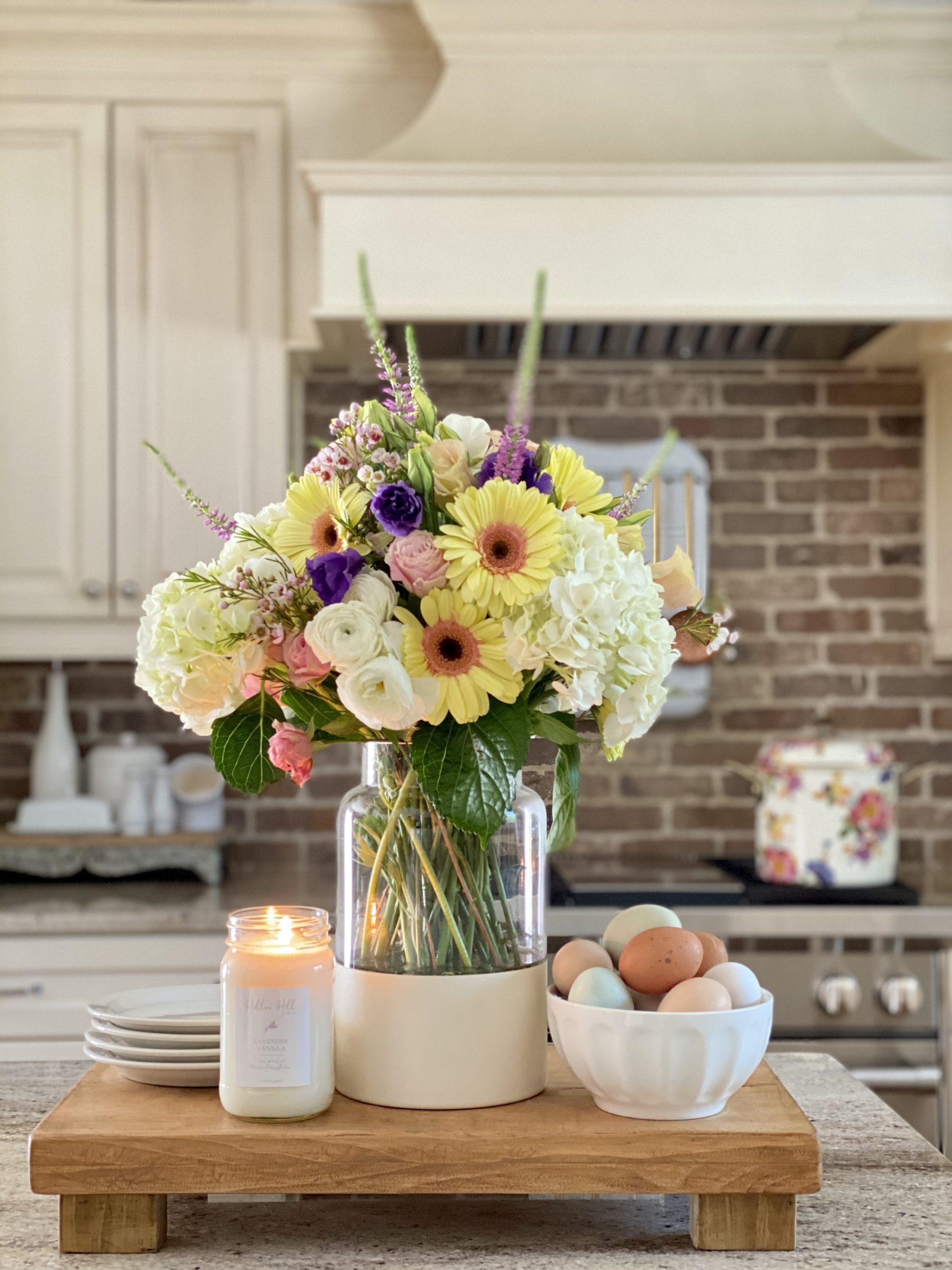 DIY color block vase on the kitchen island with a Spring floral arrangement. It is on a riser with a candle and farm fresh eggs in a white bowl.