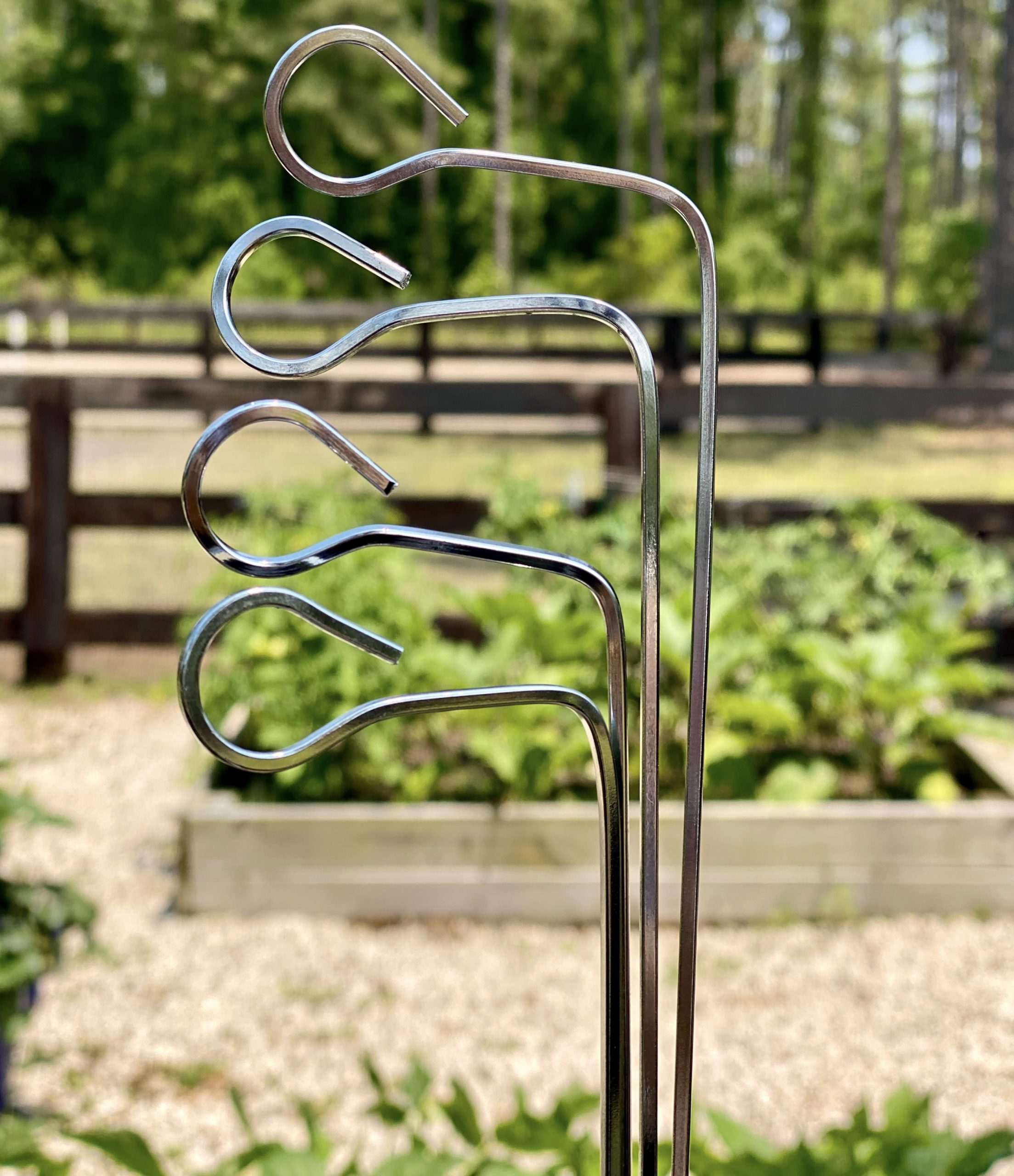 Metal skewers being held up in front of the garden. The skewers are bent at a ninety degree angle with the opening of the circle on top so that the tin can top can hang off when used as a garden marker. 