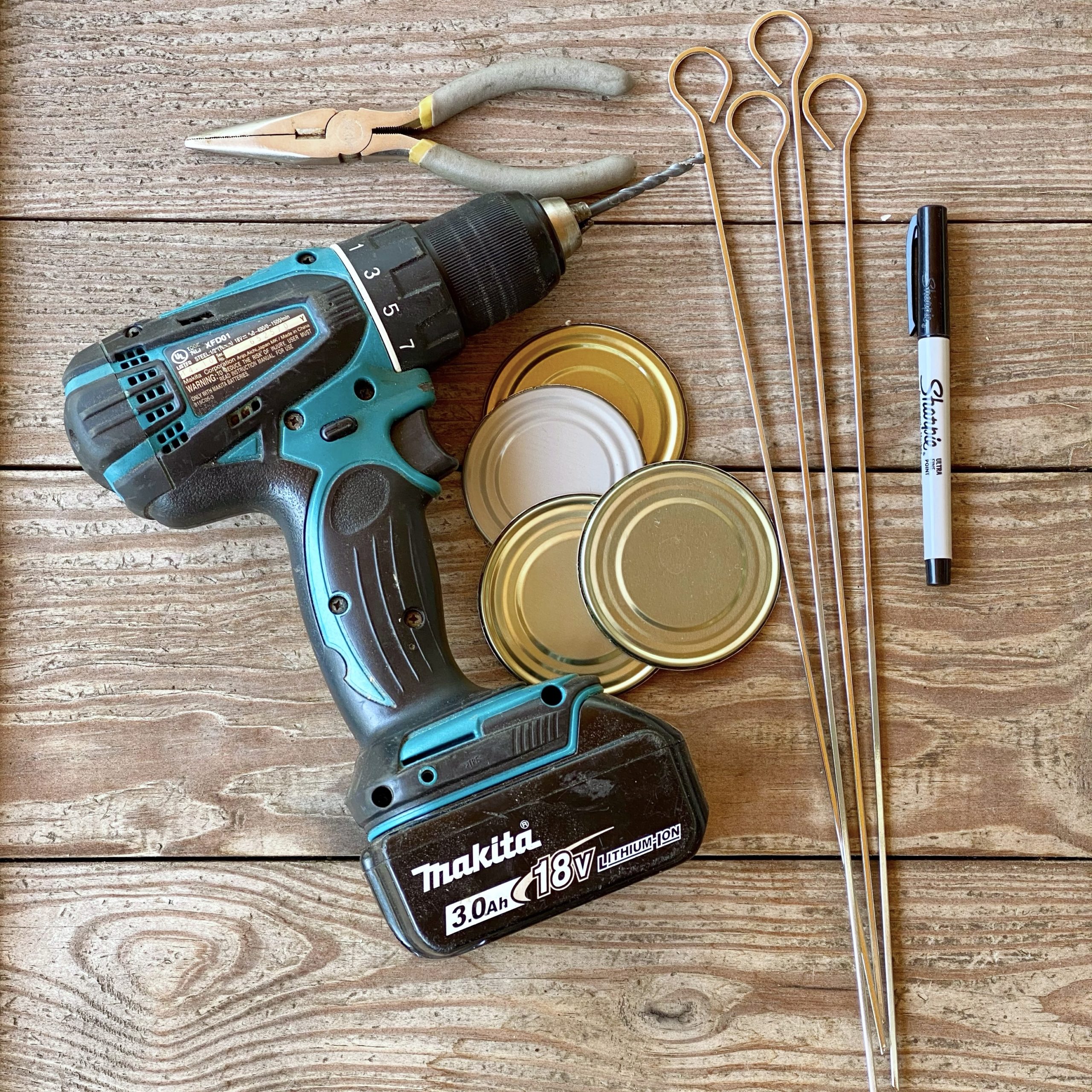 Supplies needed to make DIY garden markers: tin can tops, metal skewers, pliers, a drill, and a waterproof marker.