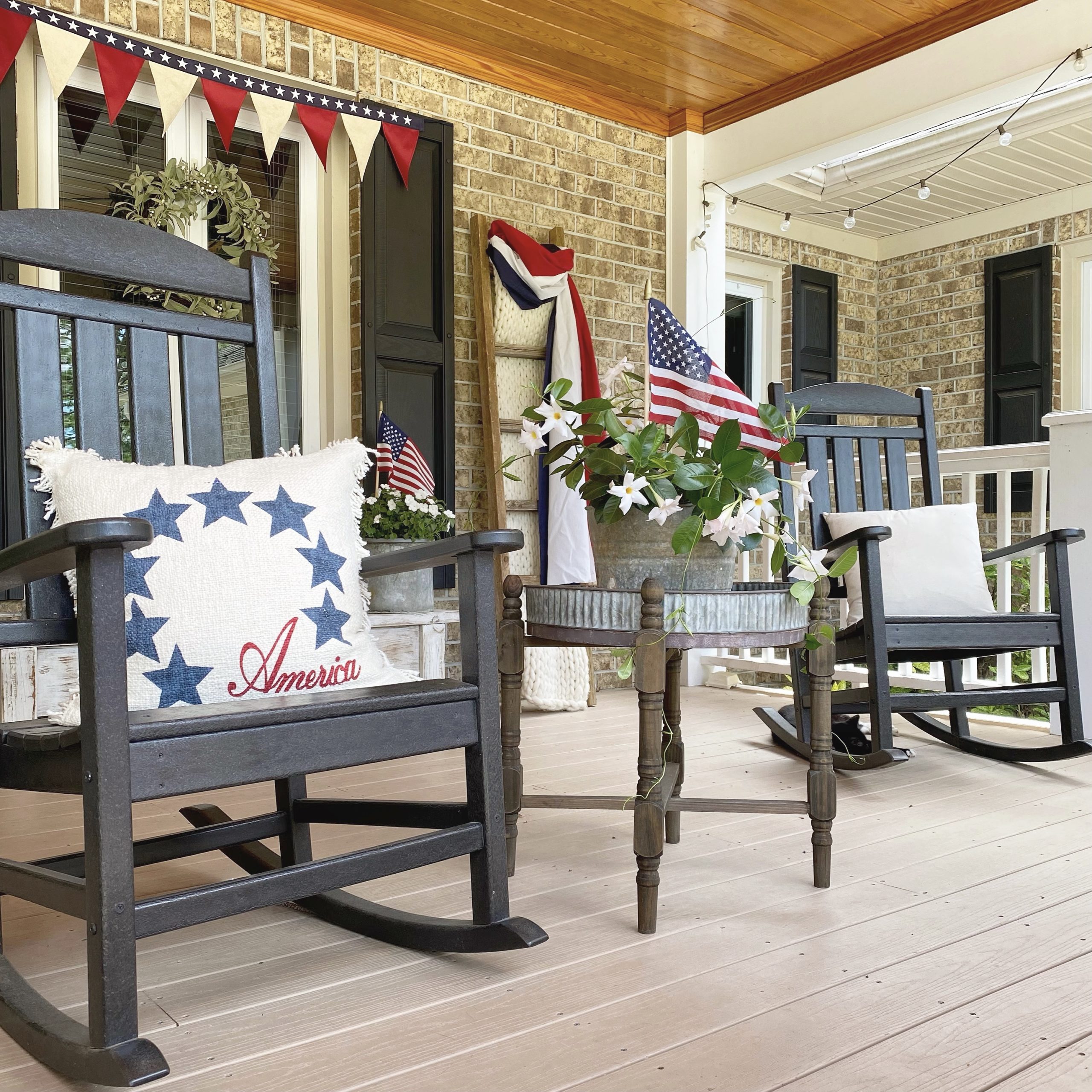 Two black rocking chairs with a table in between. There is a red, white and blue pillow on the rocker and American flags in the plant on the table. 