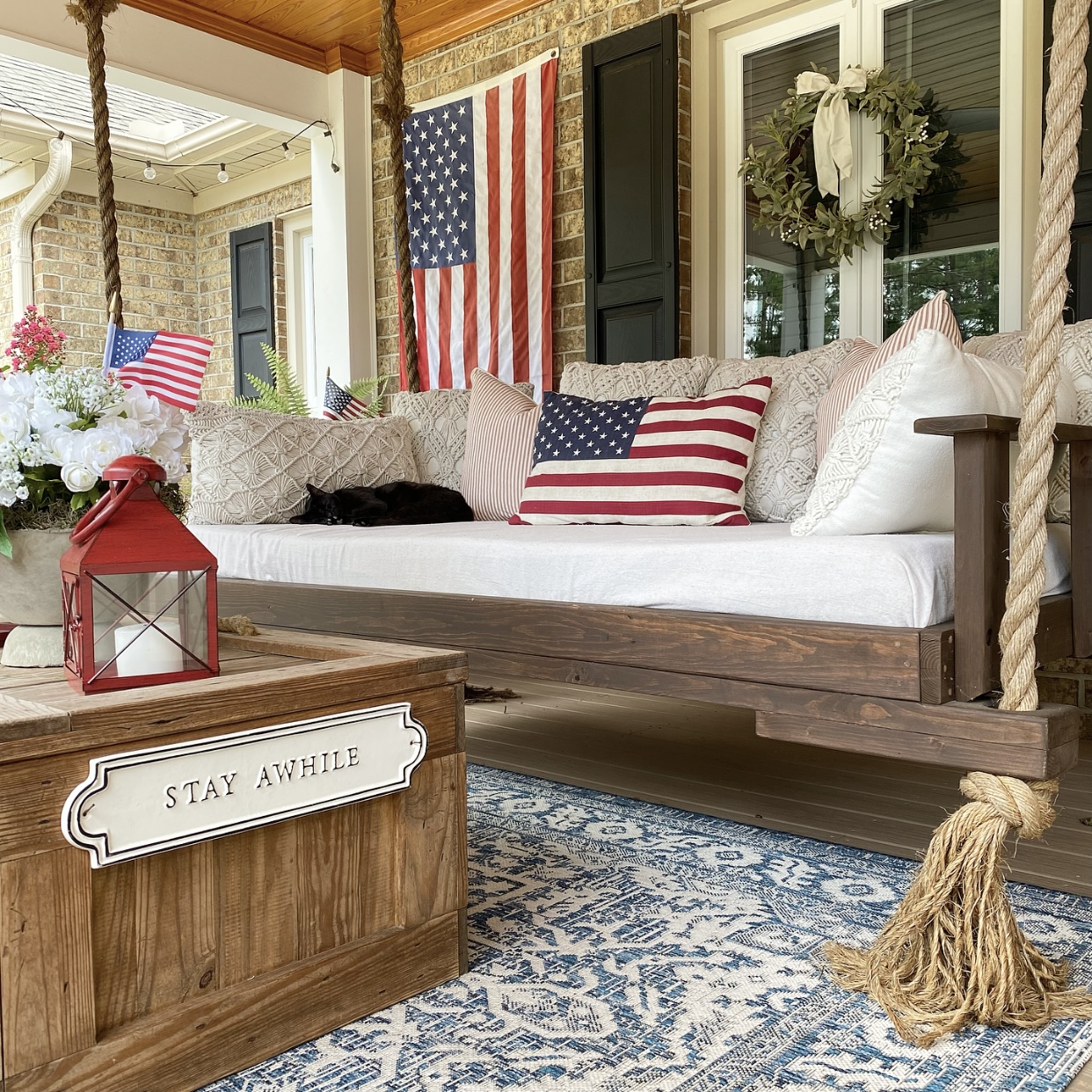 The front porch bed swing decorated with an American flag pillow. On the coffee table is a red lantern and a white floral arrangement in a concrete pedestal bowl. Behind the swing is an American flag hanging on the wall.