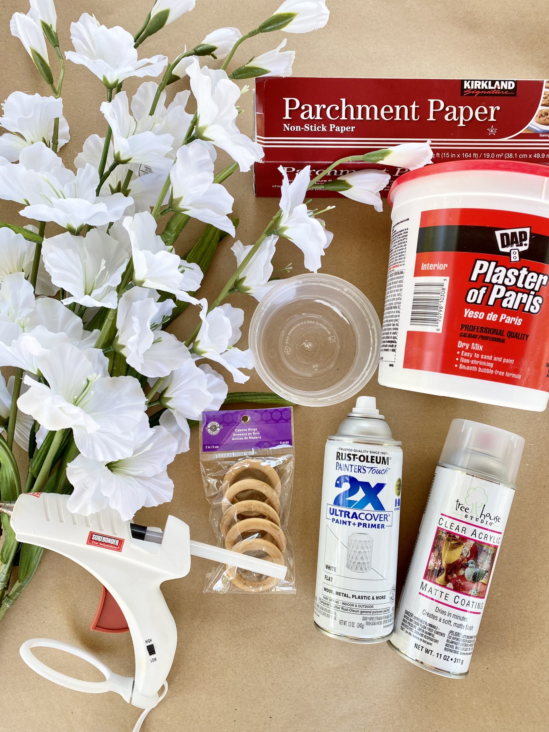 Items needed to make plaster of paris floral napkin rings including faux flowers, plaster of paris, hot glue gun, napkin rings, parchment paper, a bowl, and spray paint.
