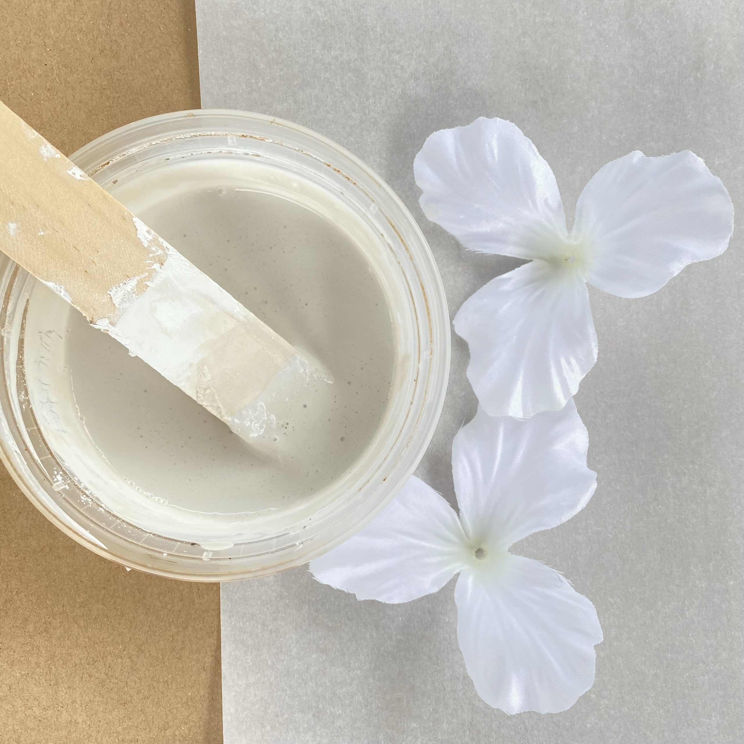 Plaster of paris in a small container with a stir stick. Next to the plaster are the petals from a white faux gladiolus flower.