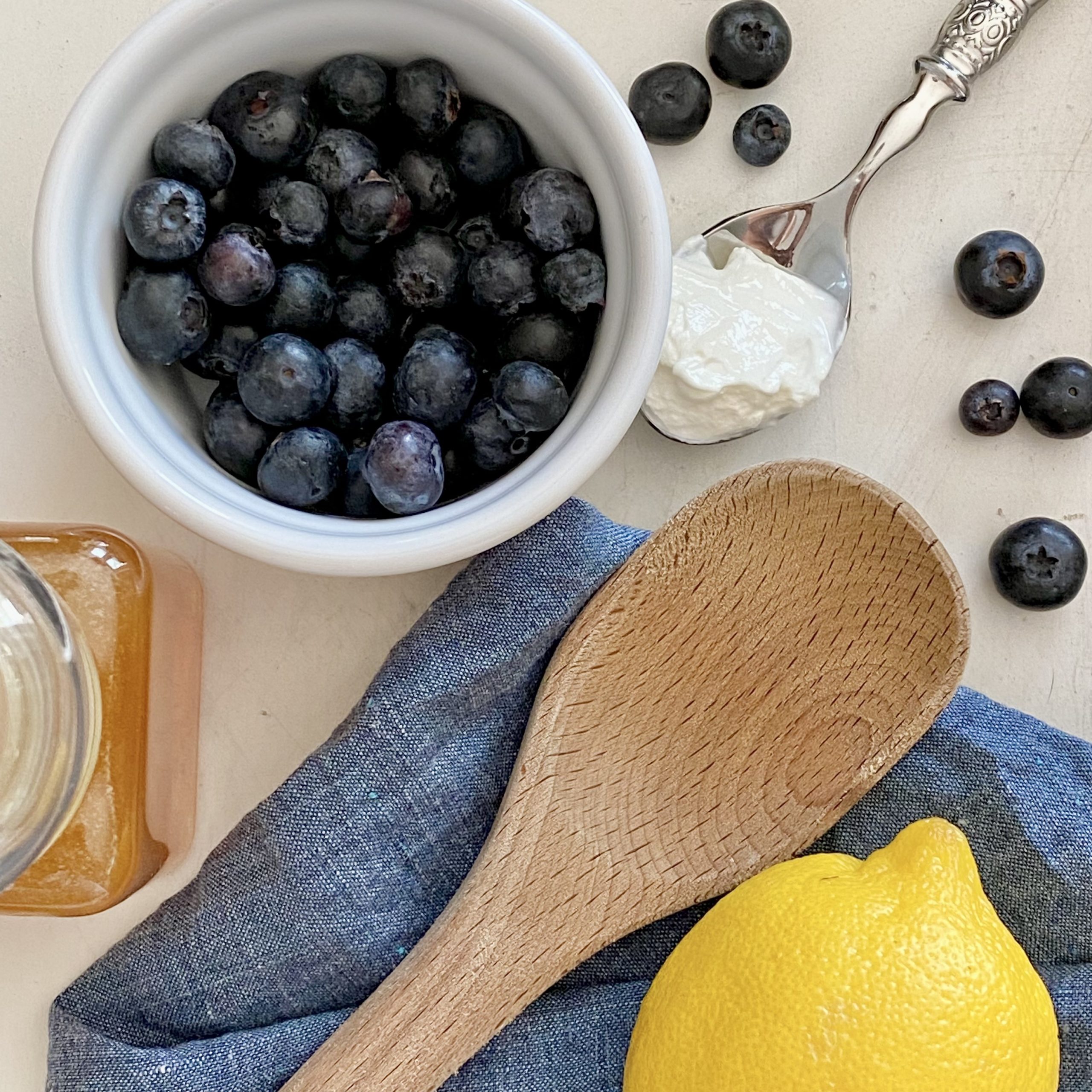 Flat-lay photo of blueberries, a lemon, honey, and yogurt with a wooden spoon and a blue towel.