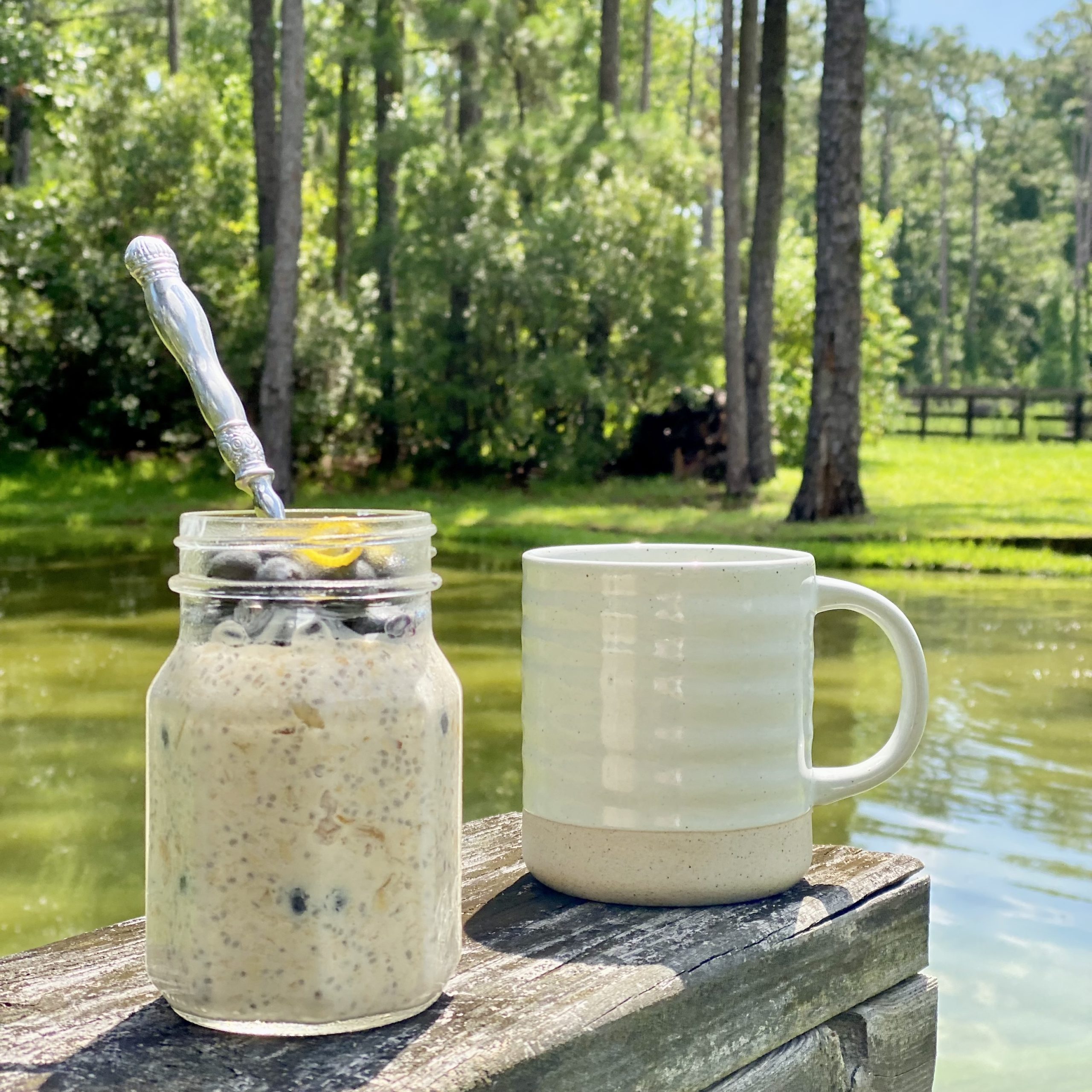 Overnight oats in a jar with a spoon in it and a coffee cup behind it next to the pond.
