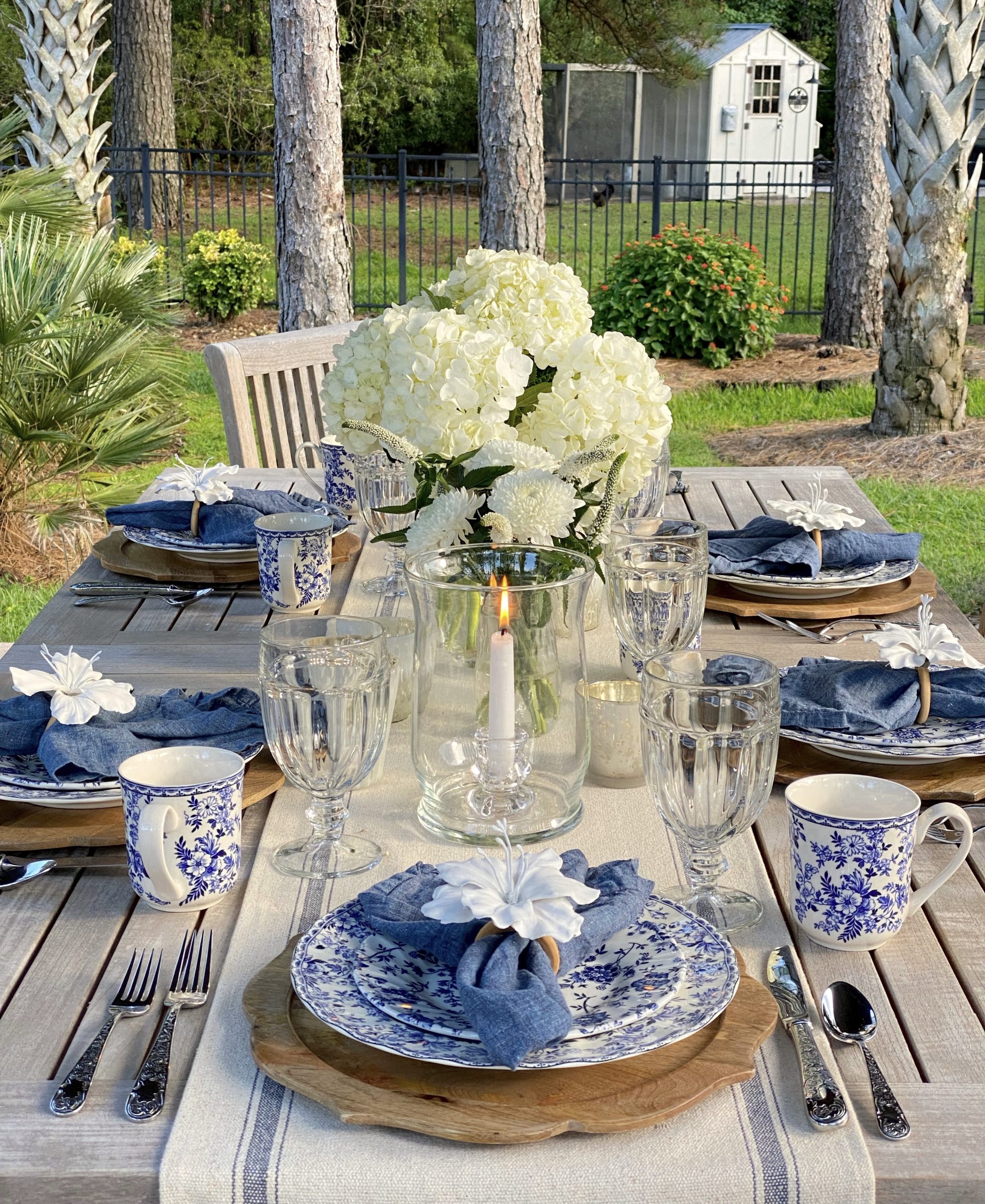 Blue and white outdoor tablescape with focus on the white floral centerpieces with chicken coop in background.