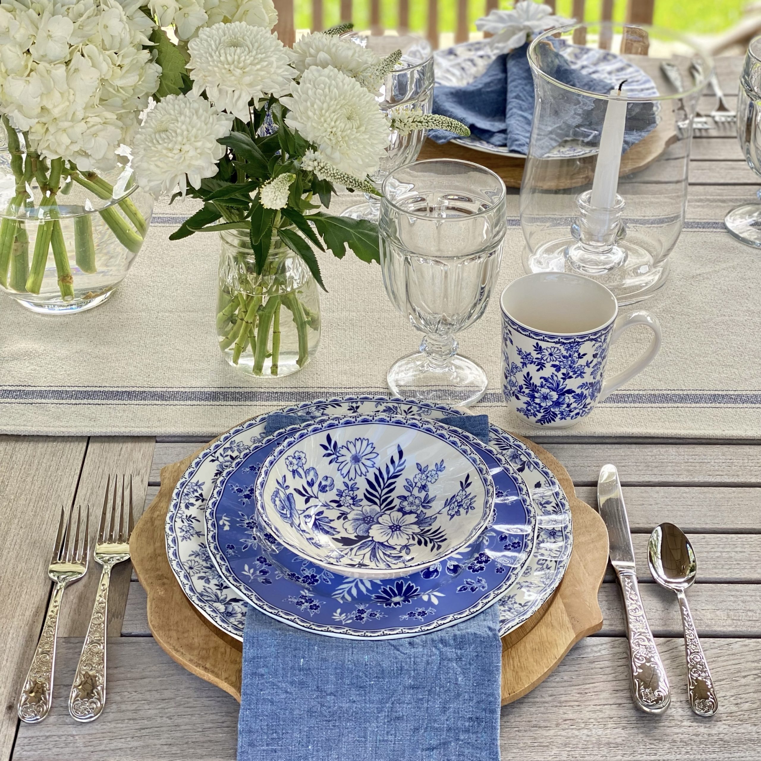 Blue and white dishes set on a wood charger with a blue linen napkin laying flat between the dinner and salad plate. There are white flowers in a jar off to the side.