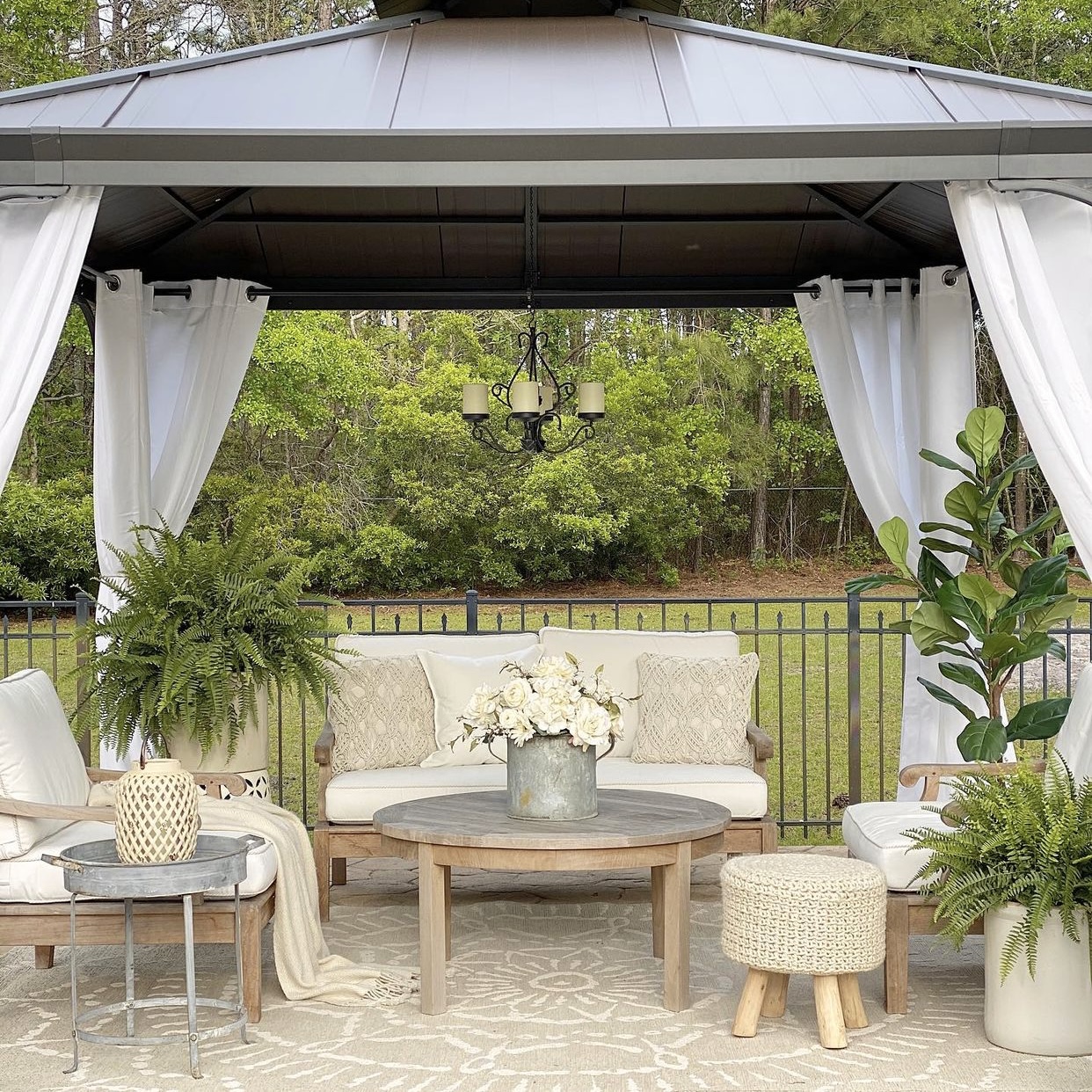 Backyard gazebo with sitting area under it. A galvanized bucket holds flowers on the coffee table and a galvanized table is next to the teak chair. 