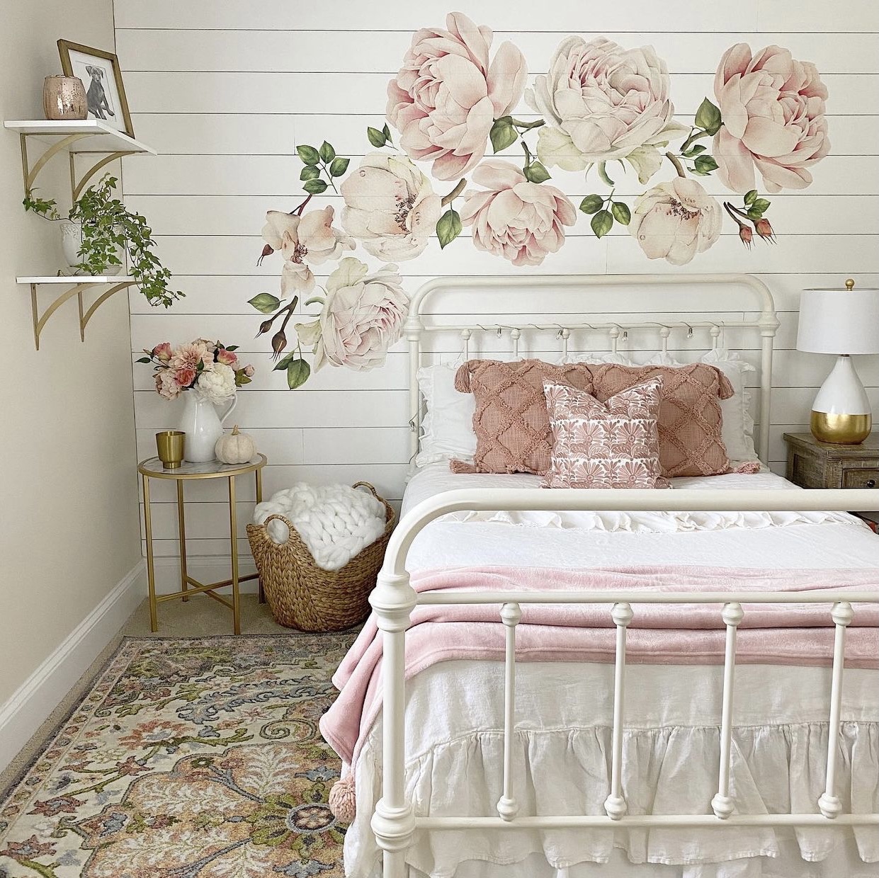 Girls bedroom with pink floral decals on white shiplap behind the white iron bed, open shelves on the opposite wall and a rug on the floor.