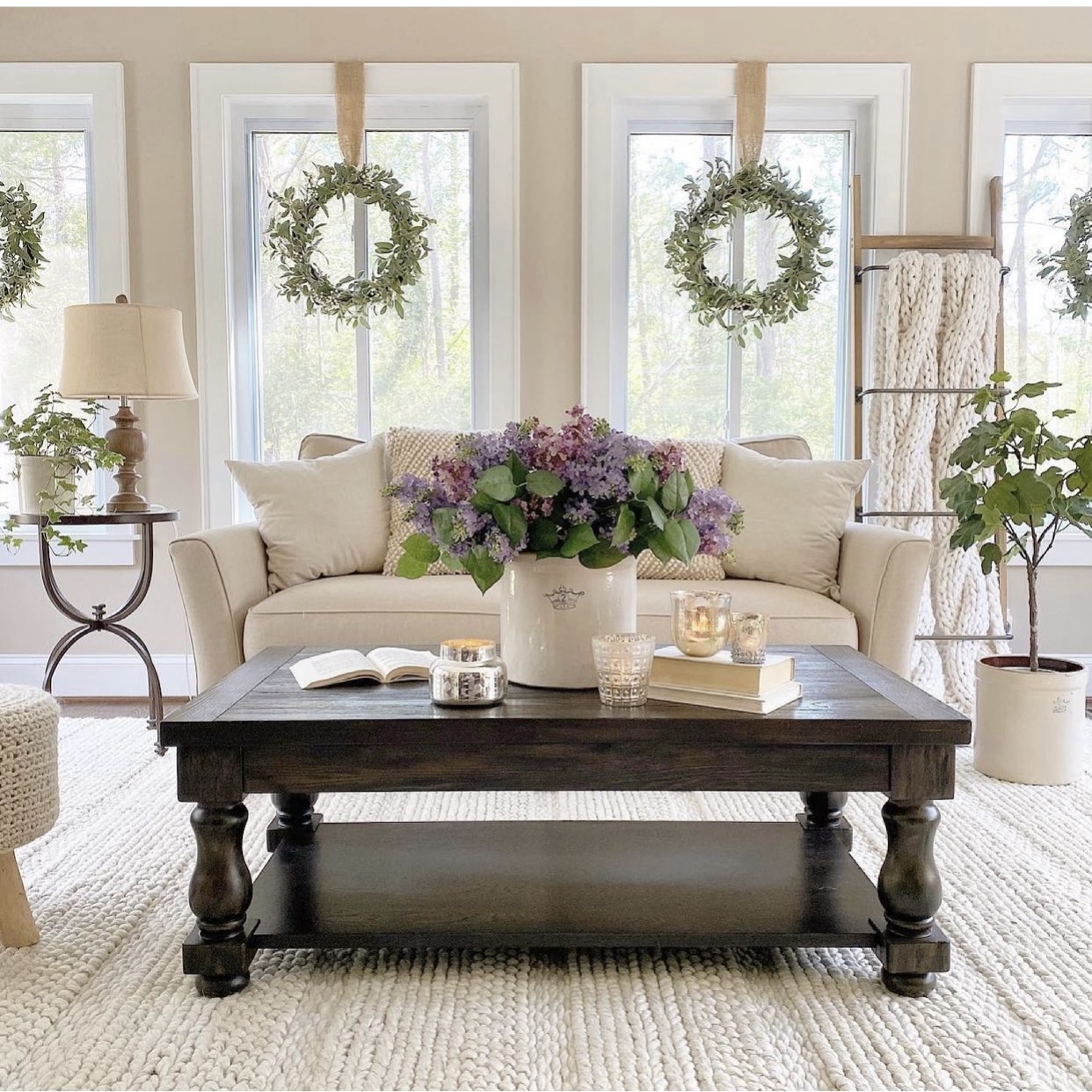 Farmhouse sunroom with a beige sofa and a coffee table in front of it with hydrangeas in a crock and candles on it. Behind the couch is a blanket ladder with a cozy blanket draped over it. 
