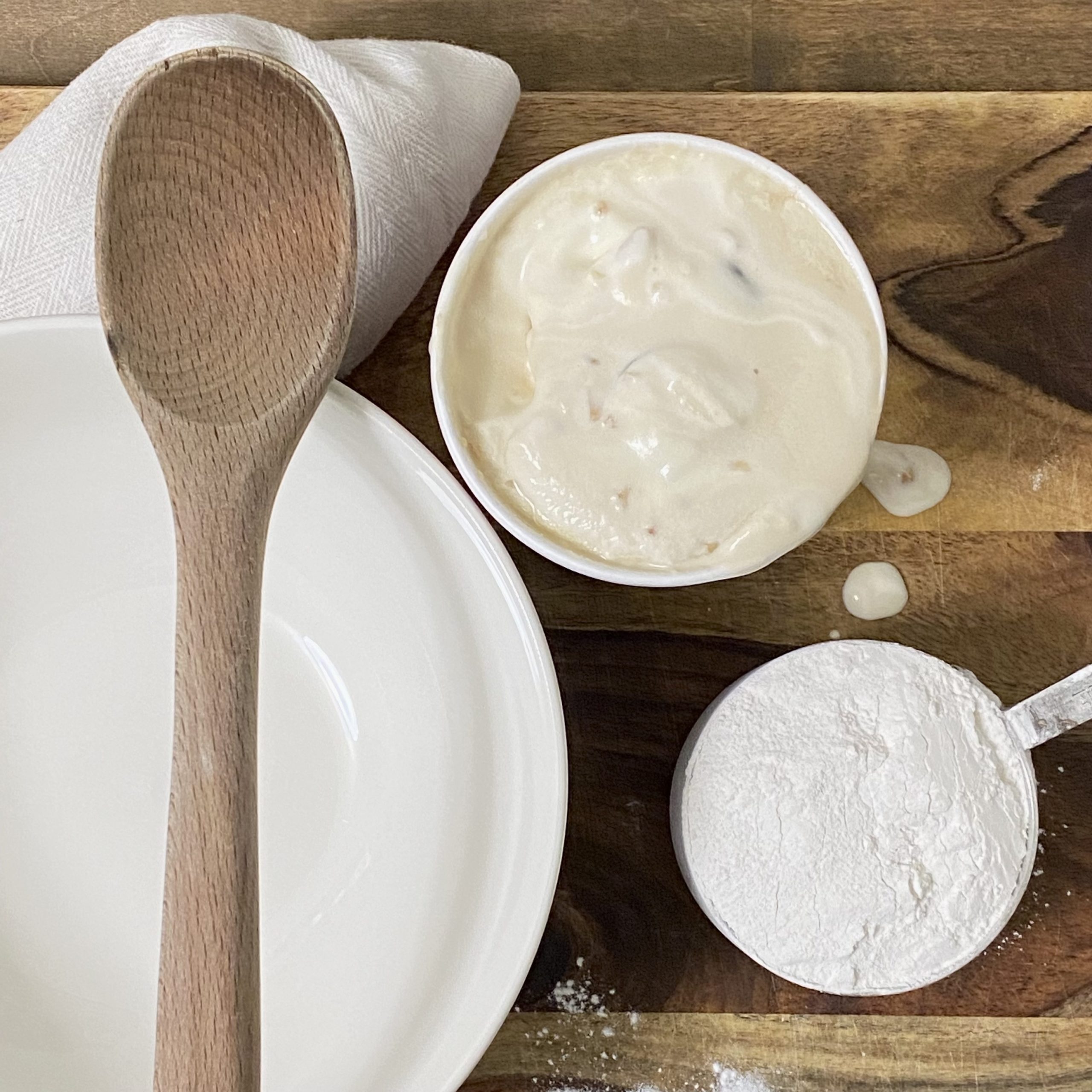 White bowl with a wooden spoon, ice cream, and flour on a wood cutting board.