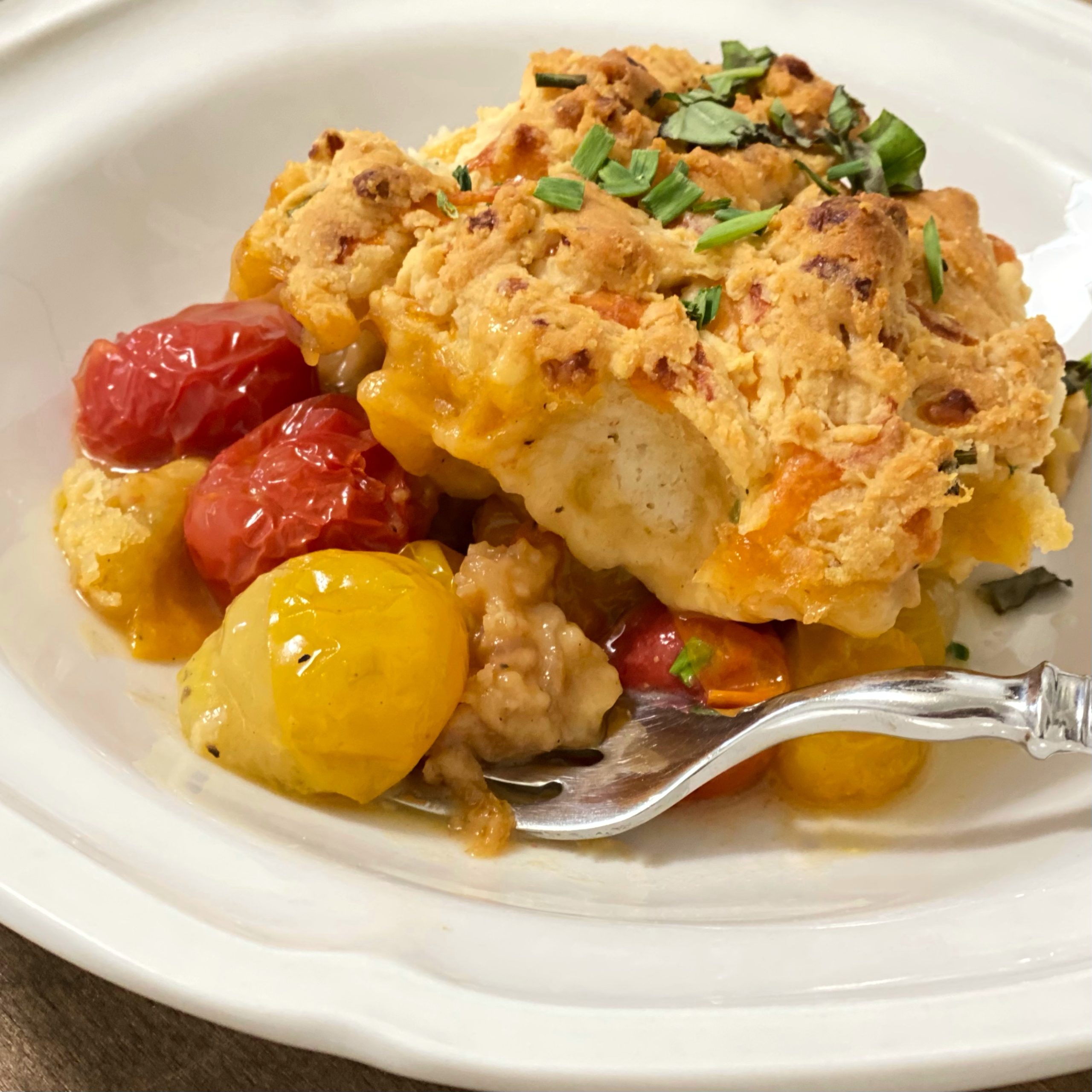 A serving of Tomato Cobbler in a white bowl with a fork. The yellow and red tomatoes look juicy and the biscuit topping is golden brown.