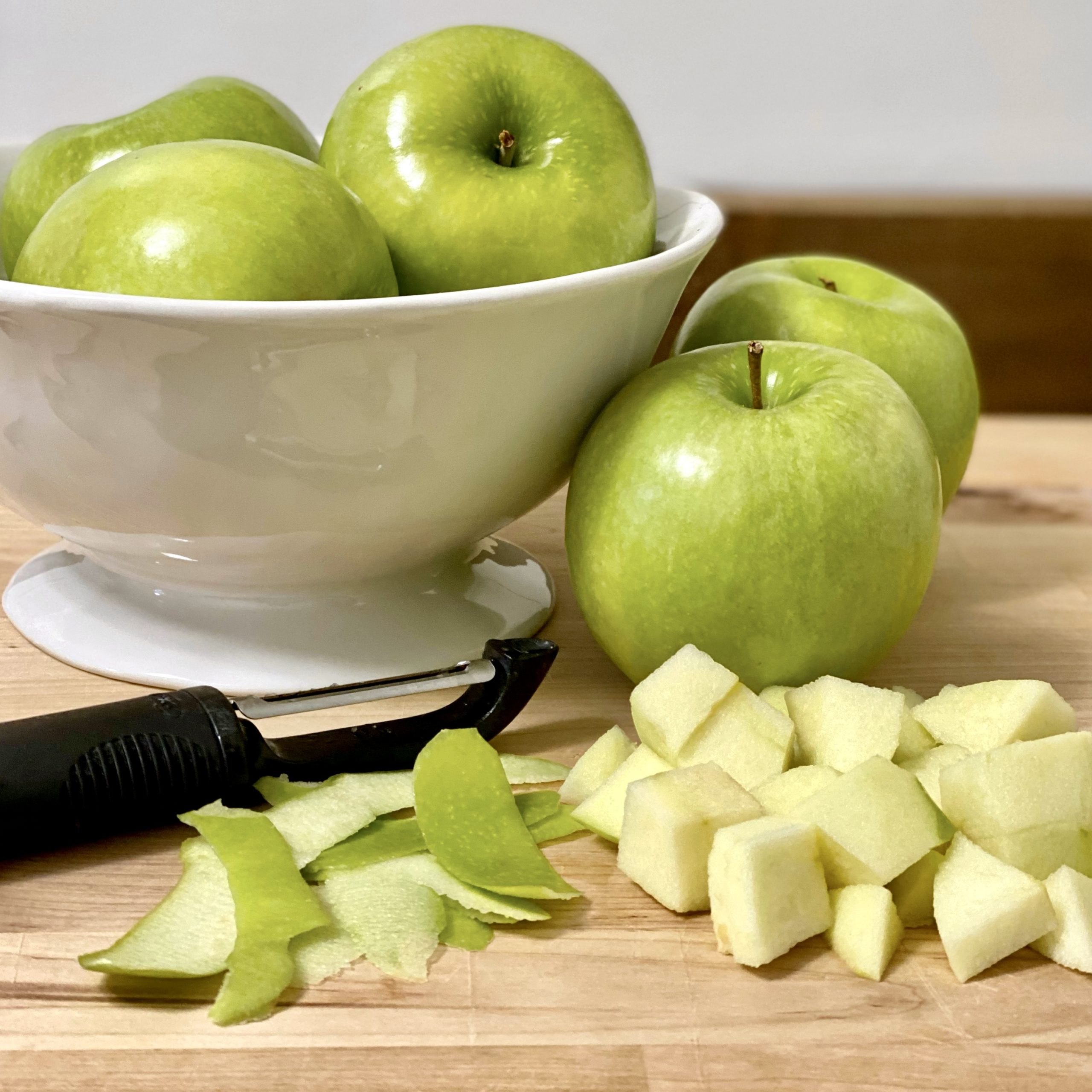 Granny Smith apples in a white pedestal bowl on a wood cutting board. More apples are on the cutting board with a peeler, apple peels, and cubed apple pieces.