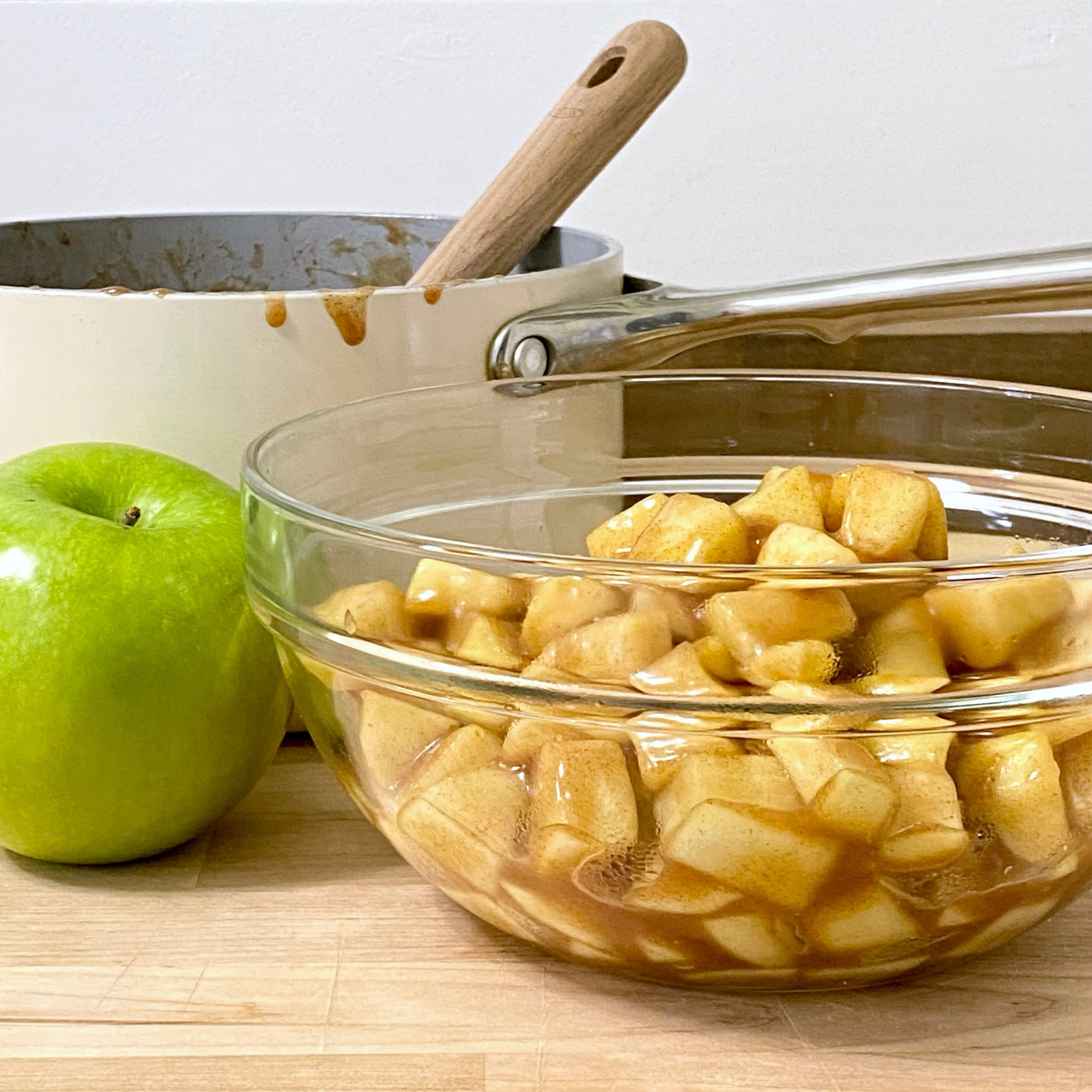 Apple pie filling in a glass bowl in from of sauce pan with a green apple next to them.