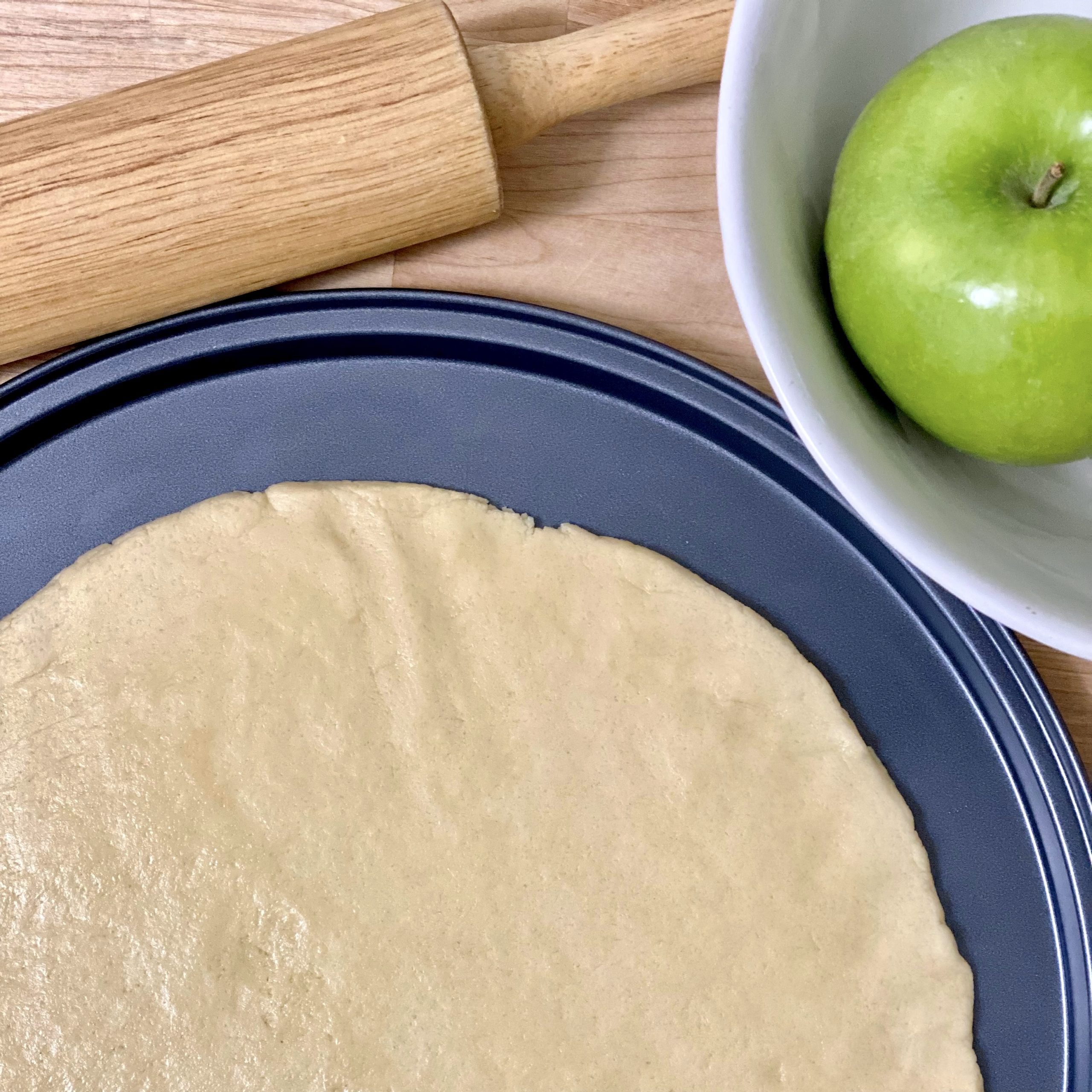 Apple pie pizza crust in the pizza pan before baking with a rolling pin and apple next to it.