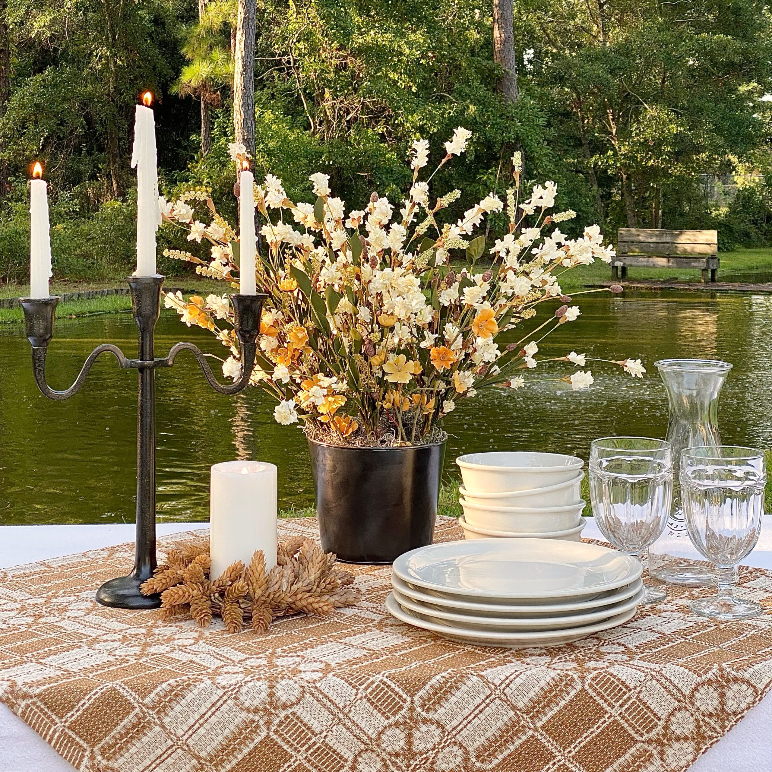 Picnic table by the pond decorated for early Fall with table cloths, candles, fall florals, plates, bowls and glasses.