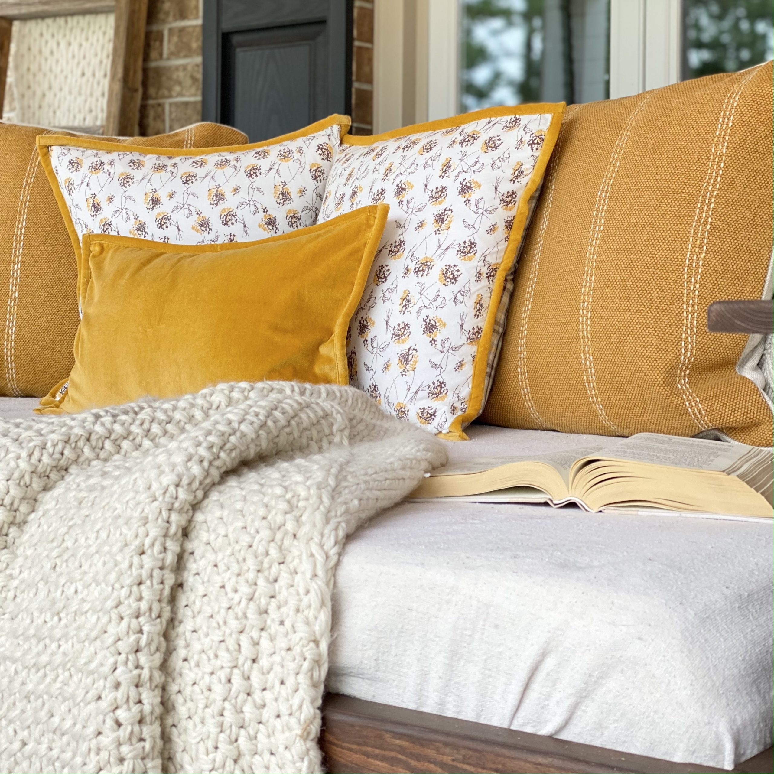 A collection of pillows on the porch swing in hues of mustard yellow, white, and cream with an off-white knit blanket and an open book.