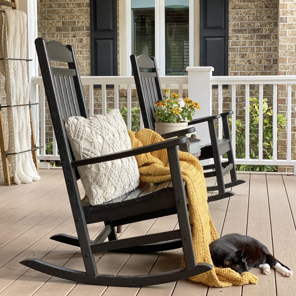Two black rocking chairs on the front porch. One has a cream macrame pillow and a muted gold knit blanket on it. In front of the rocker is a black cat napping.