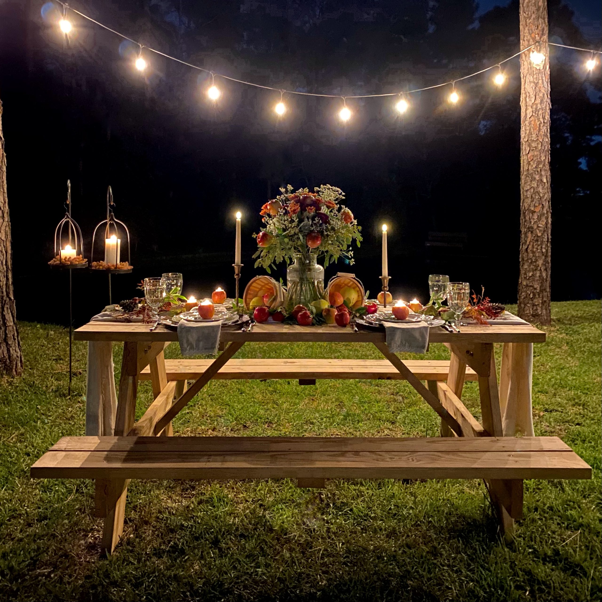 Apple harvest tablescape on a picnic table by a pond at night with white twinkle lights hanging from the trees. There is a floral arrangement, apple centerpiece, and apple votive candle holders at each place setting.