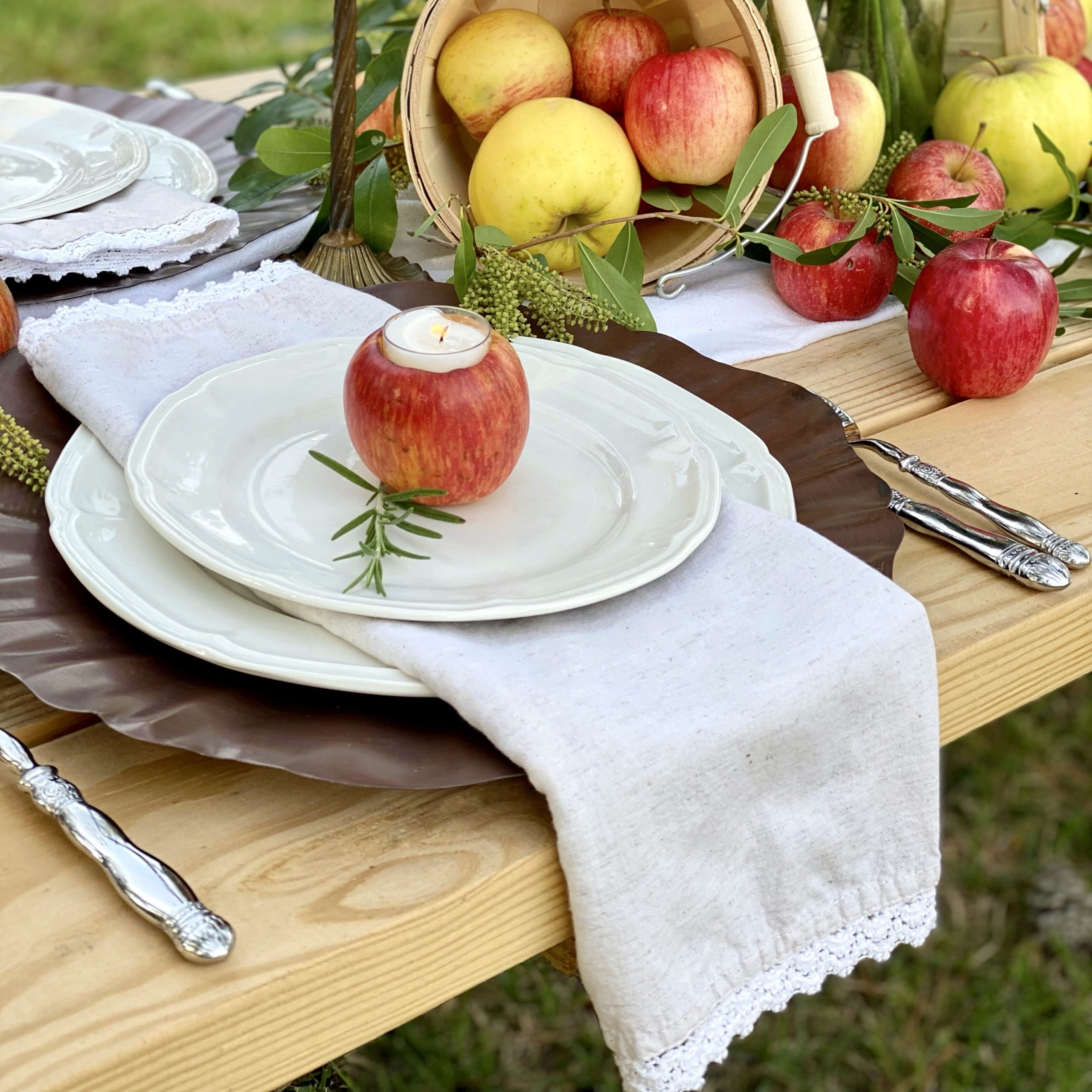 Place setting with an antique copper charger and white dishes on top. There is a linen napkin folded in thirds between the dinner and salad plate and an apple votive holder and a rosemary sprig are on the salad plate..