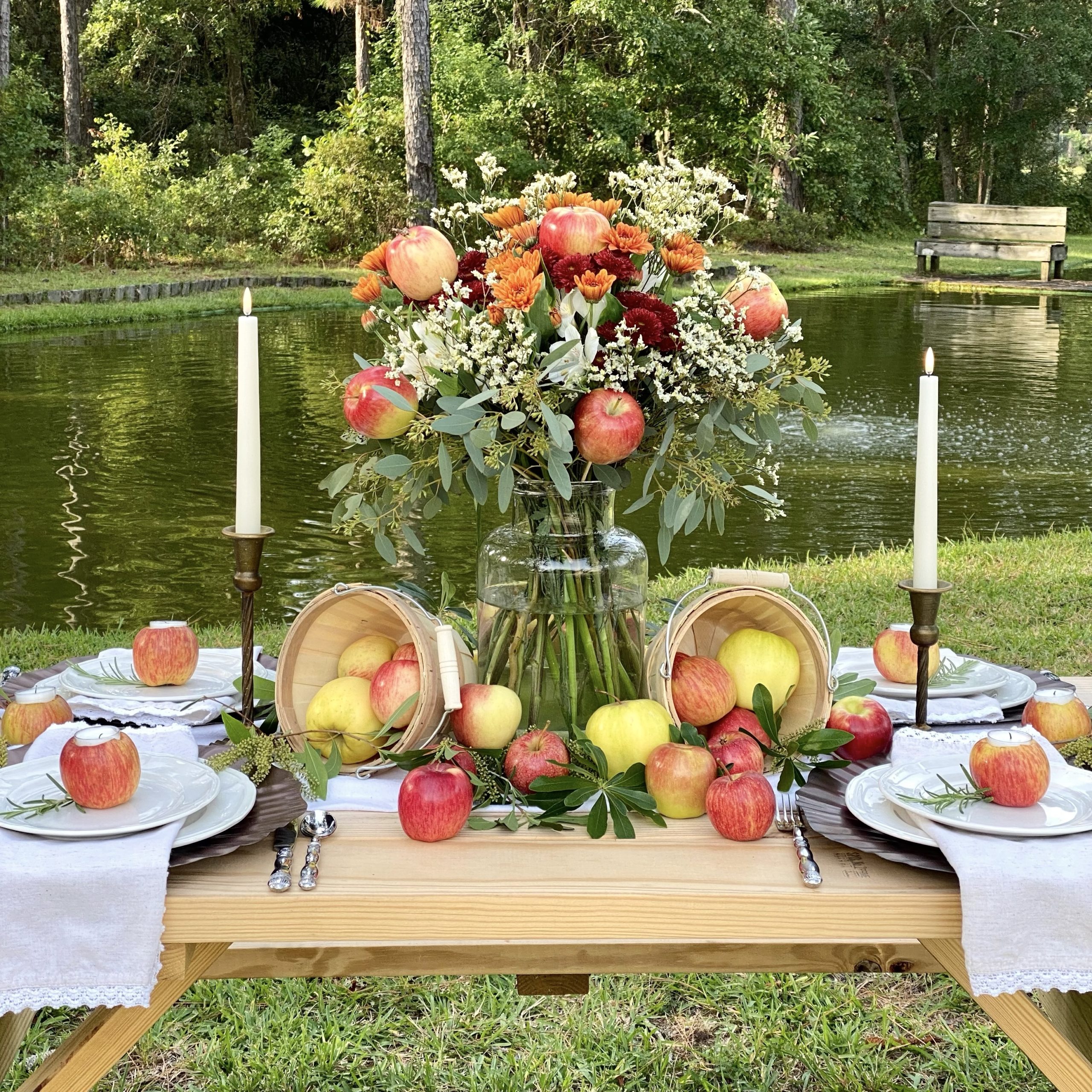 Apple harvest tablescape on a picnic table by a pond. There is a floral arrangement, apple centerpiece, and apple votive candle holders at each place setting.