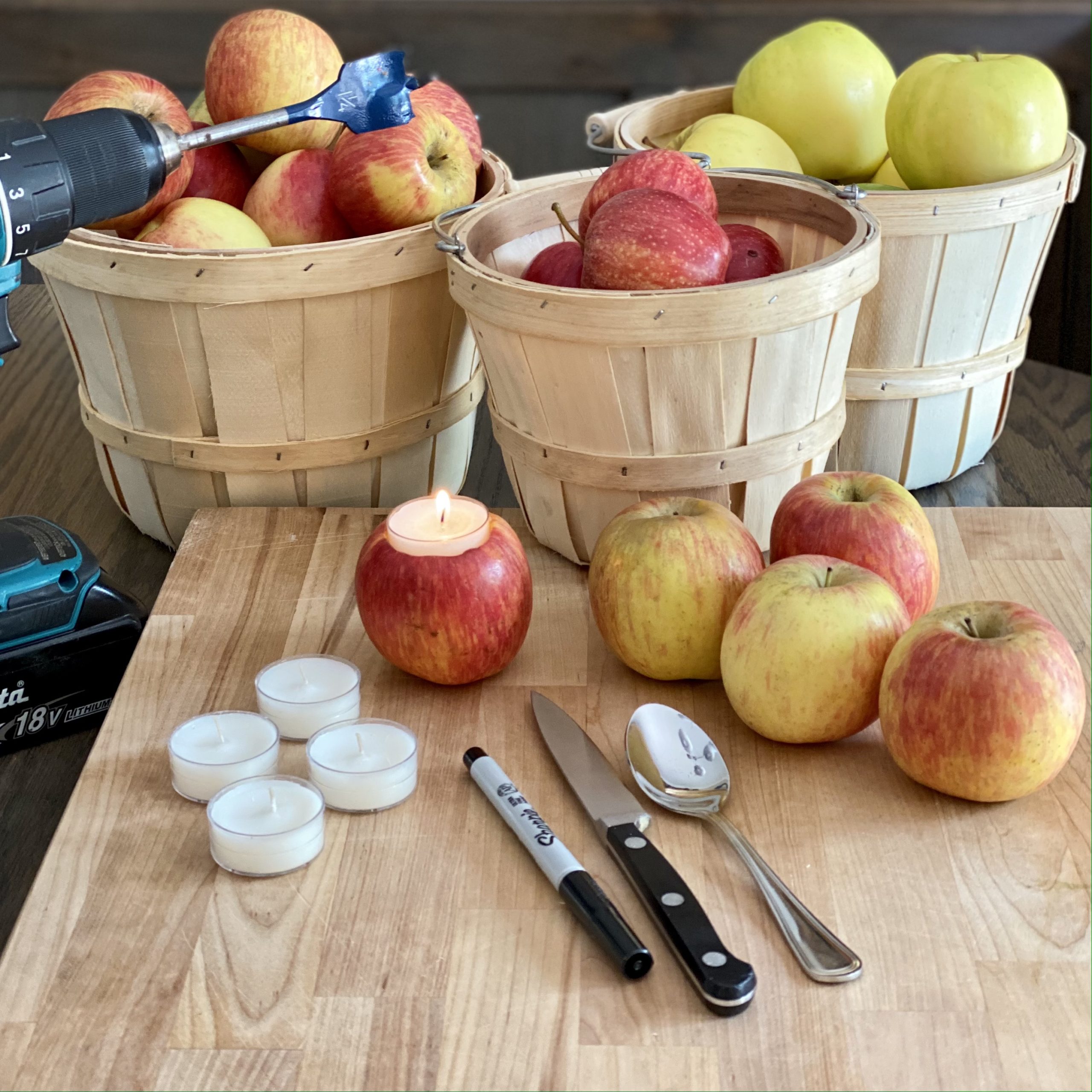 Orchard baskets filled with apples in the background. In the foreground is everything needed to make apple votives including apples, votive candles, a paring knife, spoon, Sharpie, and a drill with a spade bit on it.