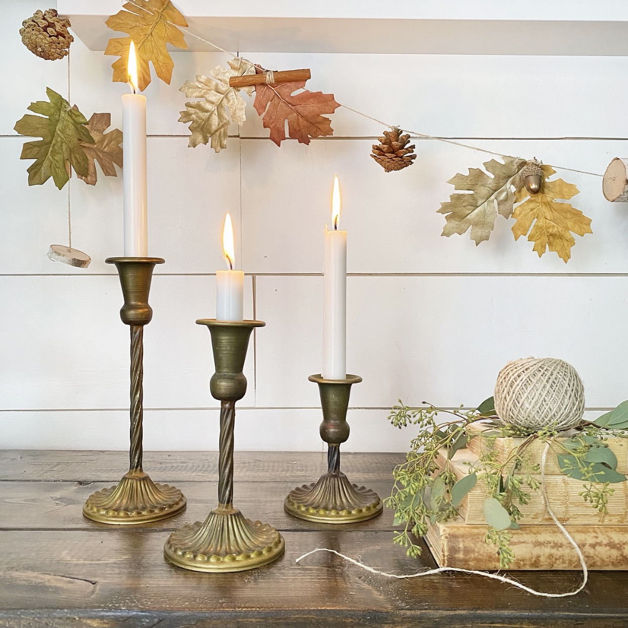 Easy Fall garland hanging from a shelf with three antique brass candlesticks with lit candles below it. 