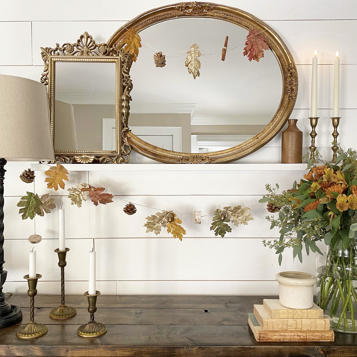 Table with antique bronze candlesticks, Fall florals, and old books styled on it. A shelf on the wall above the table has layered mirrors and Fall garland hanging from it.