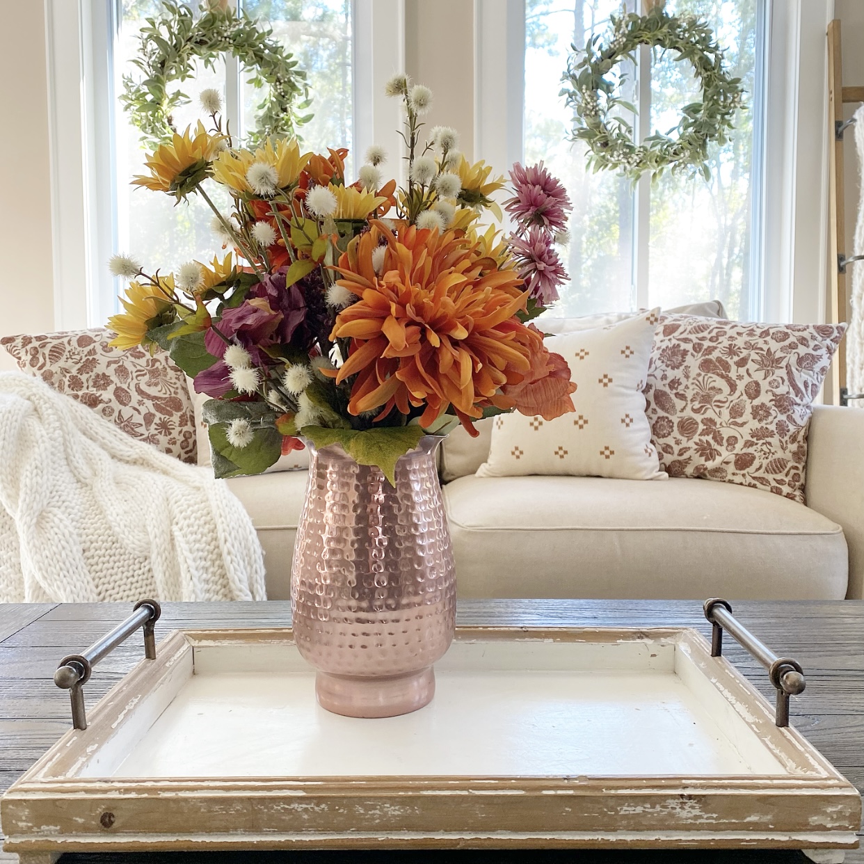 Copper pitcher filled with Fall florals on a tray on a coffee table. In the background is a sofa with Fall pillows on it and a cozy knit blanket. There are wreaths hanging from the windows.