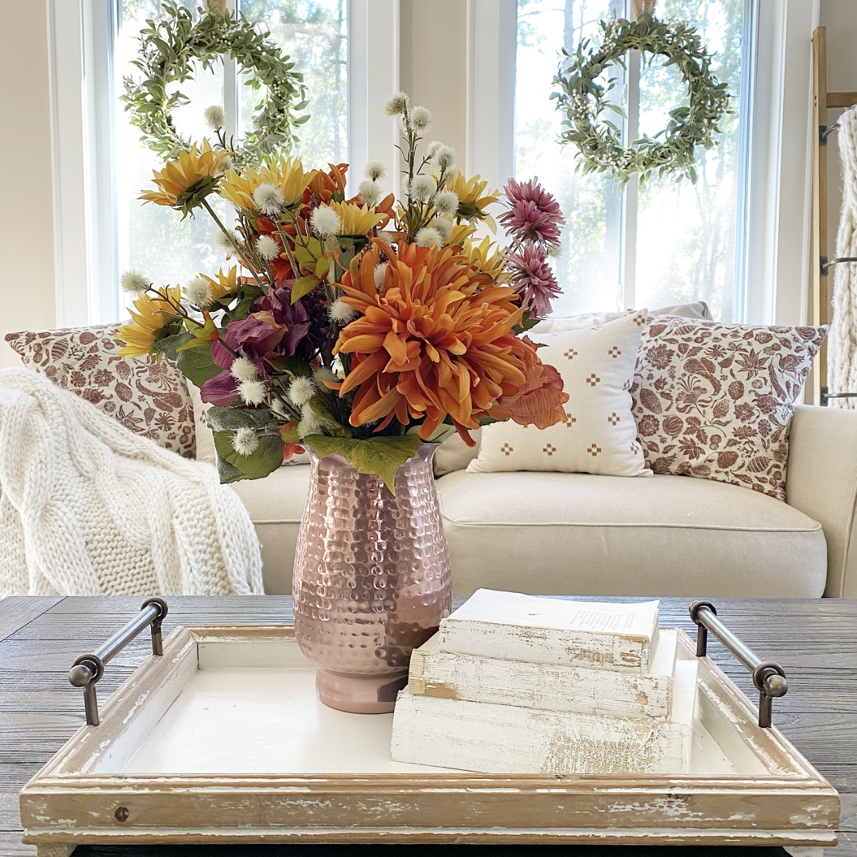 Copper pitcher filled with Fall florals and some vintage books stacked upon a tray on a coffee table. In the background is a sofa with Fall pillows on it and a cozy knit blanket. There are wreaths hanging from the windows.