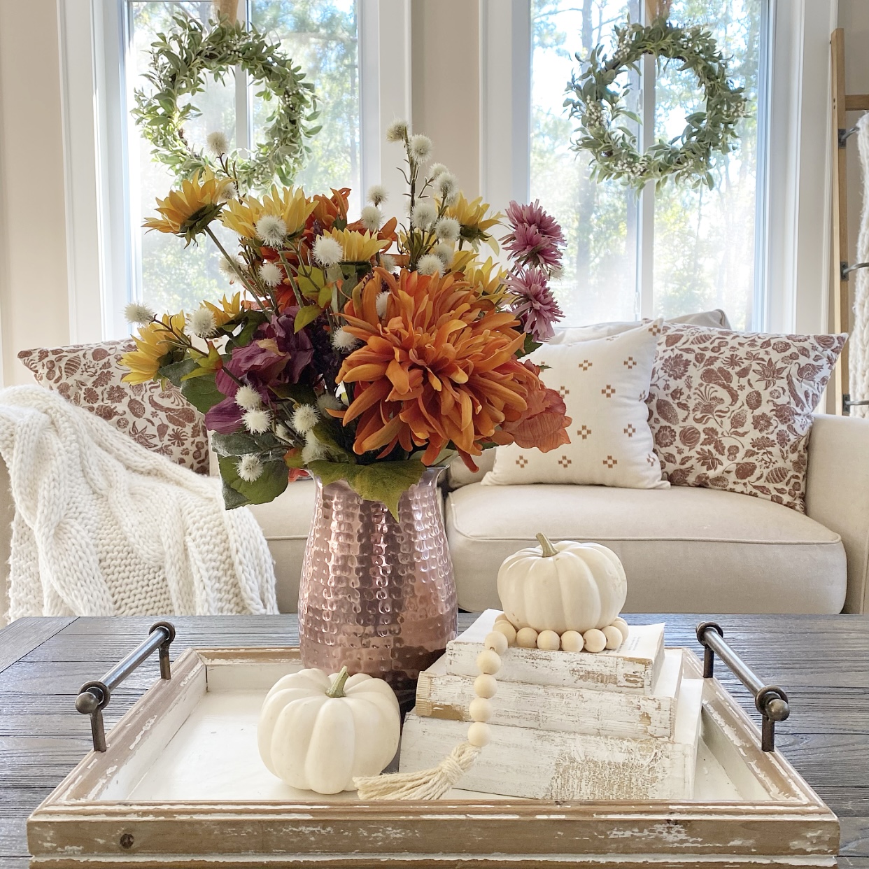 Copper pitcher filled with Fall florals, vintage stacked books, white mini pumpkins, and wood beads on a tray on a coffee table. In the background is a sofa with Fall pillows on it and a cozy knit blanket. There are wreaths hanging from the windows.