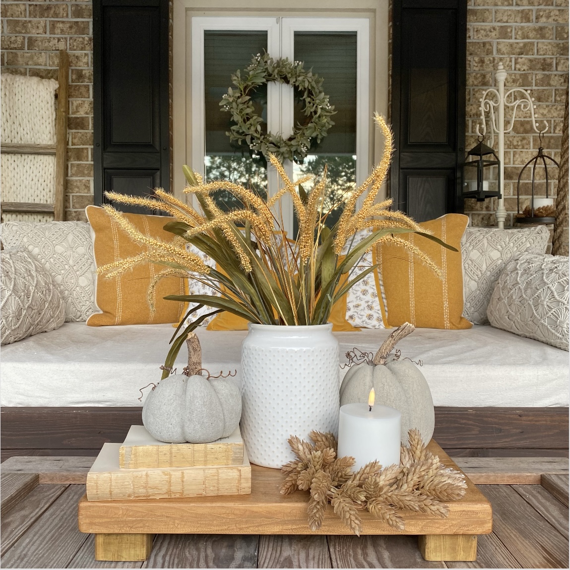 Fall vignette on the a coffee table on the front porch. The vignette includes Fall florals in a white crock, concrete pumpkins, a candle and vintage books stacked on a riser.