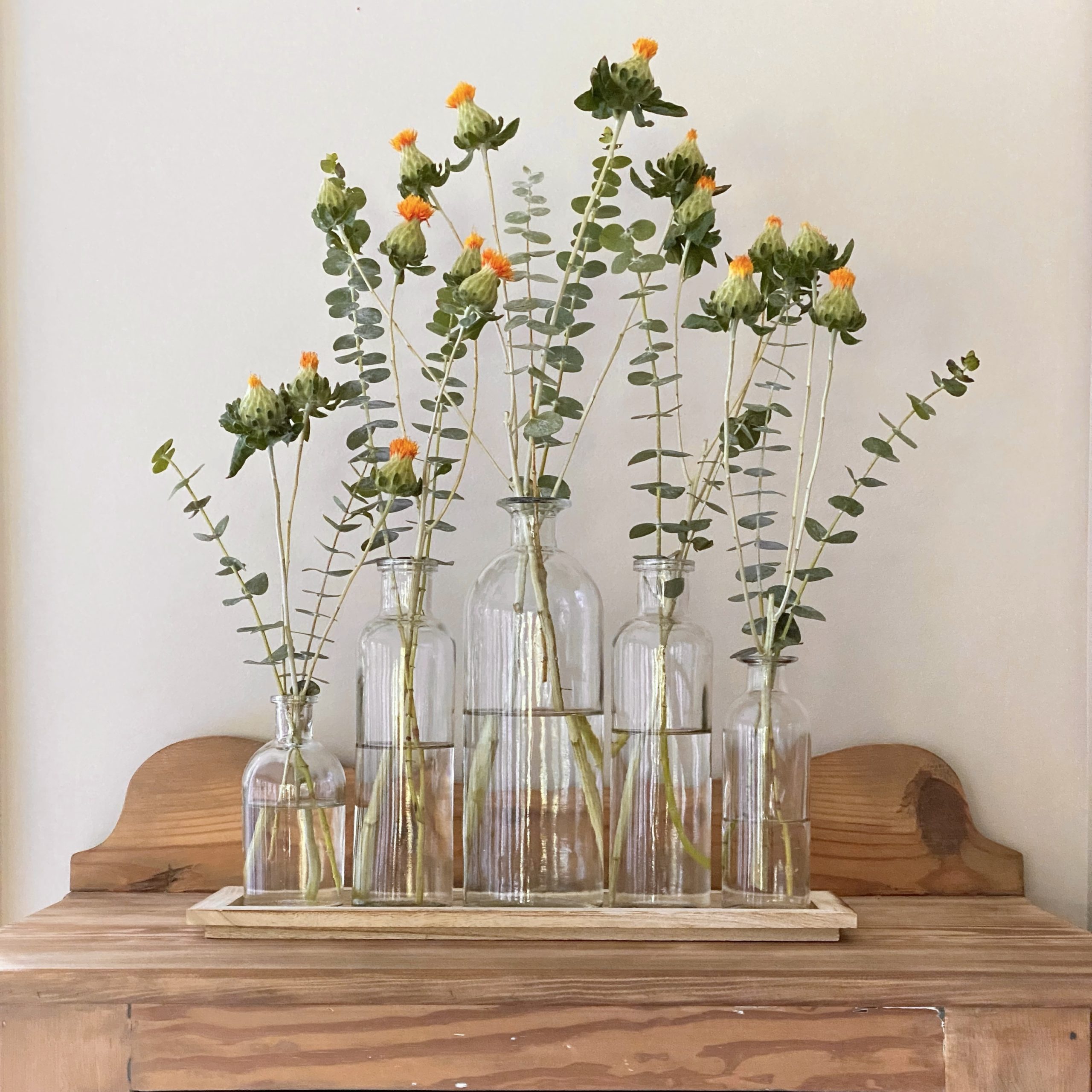 Vase set with 5 bottles on a wood tray with flowers in them on a table.
