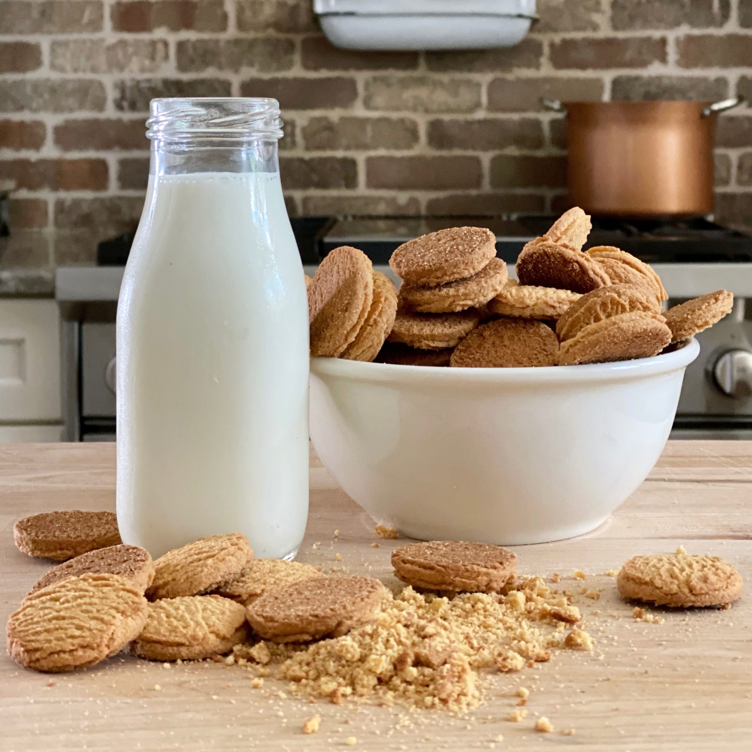 Gingersnap cookies in a white bowl with a glass jug of milk. In front of them are more gingersnap cookies, whole and crushed.