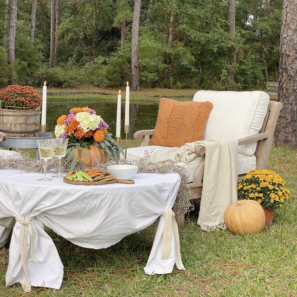 View of cozy chair with a blanket and pillows on it. In the background is the pond. In the foreground is a table with a fall floral arrangement, snacks, and candles.