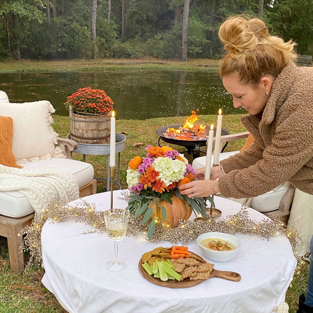 Me arranging fall florals on the table by the pond. A bonfire is lit, wine is poured and snacks are out.