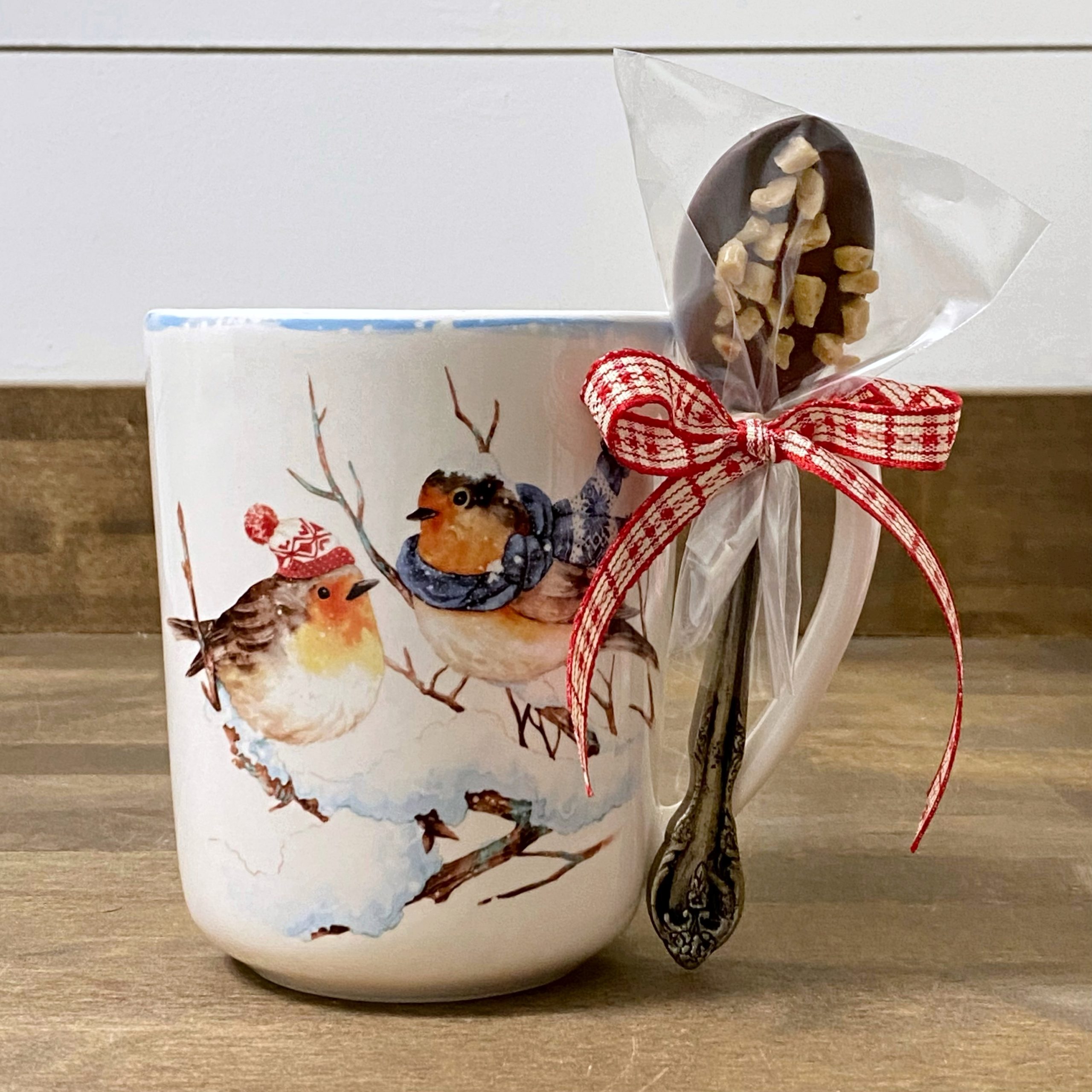 A chocolate dipped spoon tied onto the handle of a mug with winter birds on it.