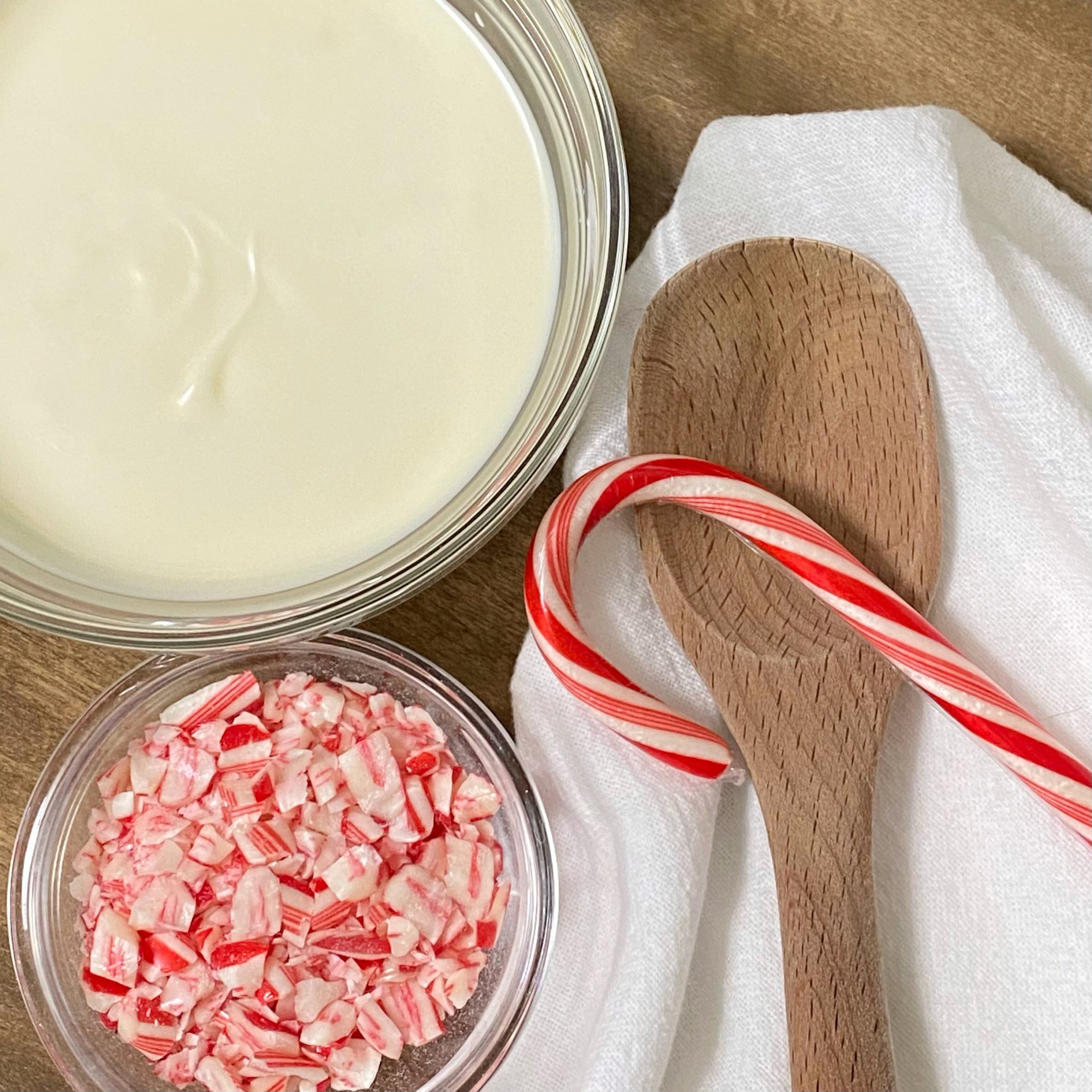 White chocolate melted in a glass bowl with crushed candy cane in another glass bowl next to it. Beside them are a white towel and wooden spoon.